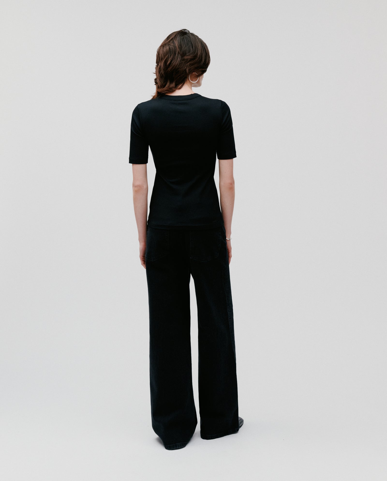 A person stands facing away, wearing the black KRISTIN Top by IVY OAK in organic cotton with wide-leg black pants against a plain background.