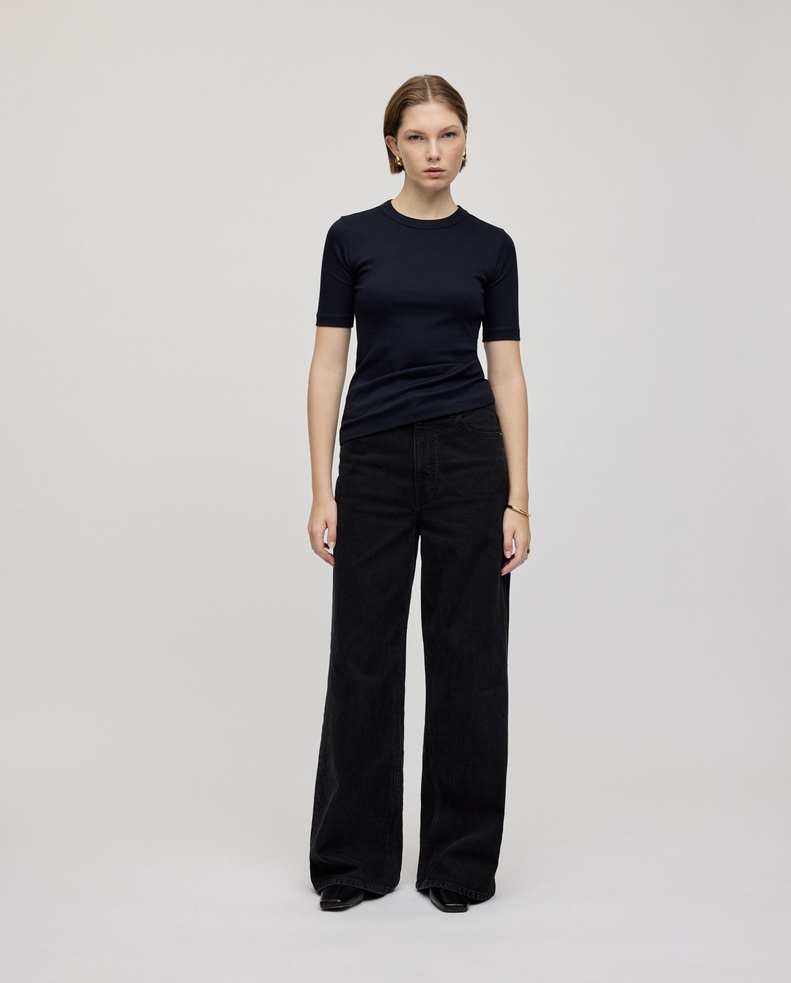 A woman stands against a plain background, wearing the IVY OAK KRISTIN Top—a comfortable, fitted black T-shirt crafted from organic cotton—paired with wide-leg black jeans.