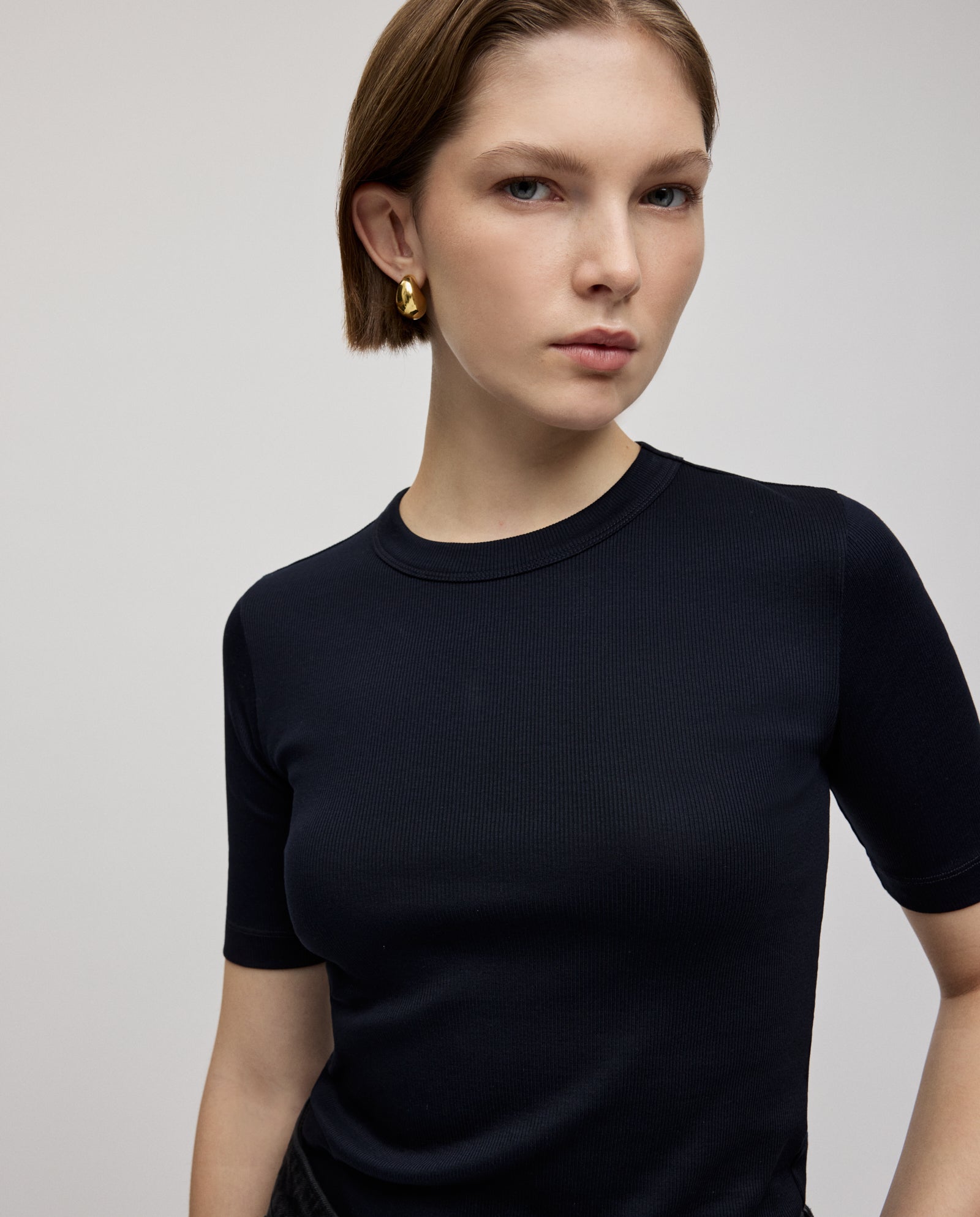 A woman with short brown hair confidently looks at the camera, wearing the KRISTIN Top by IVY OAK in black organic cotton, paired with gold hoop earrings.