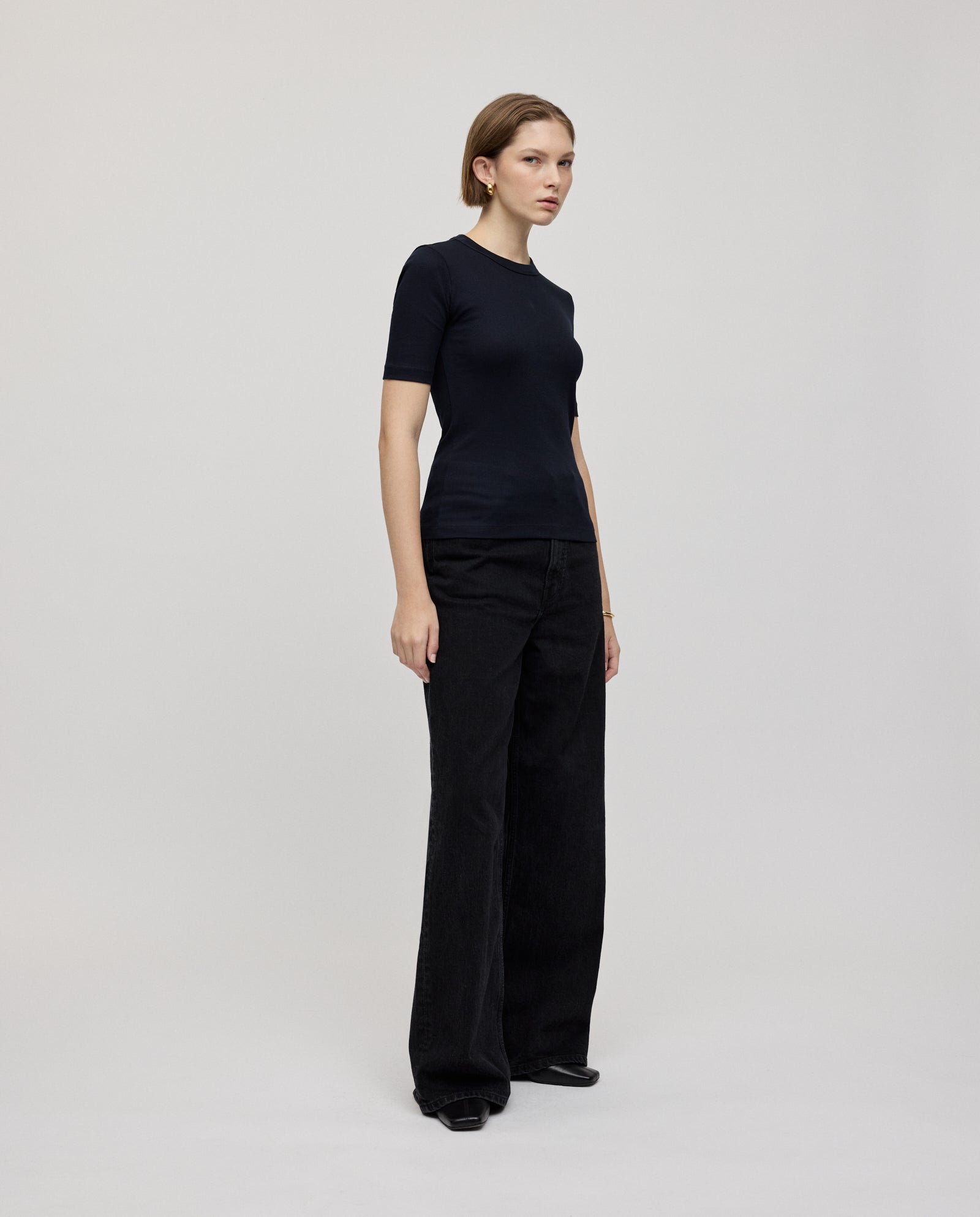 A woman stands in a studio against a plain background, wearing the KRISTIN Top by IVY OAK—an elegant, fitted black organic cotton t-shirt paired with loose black pants.
