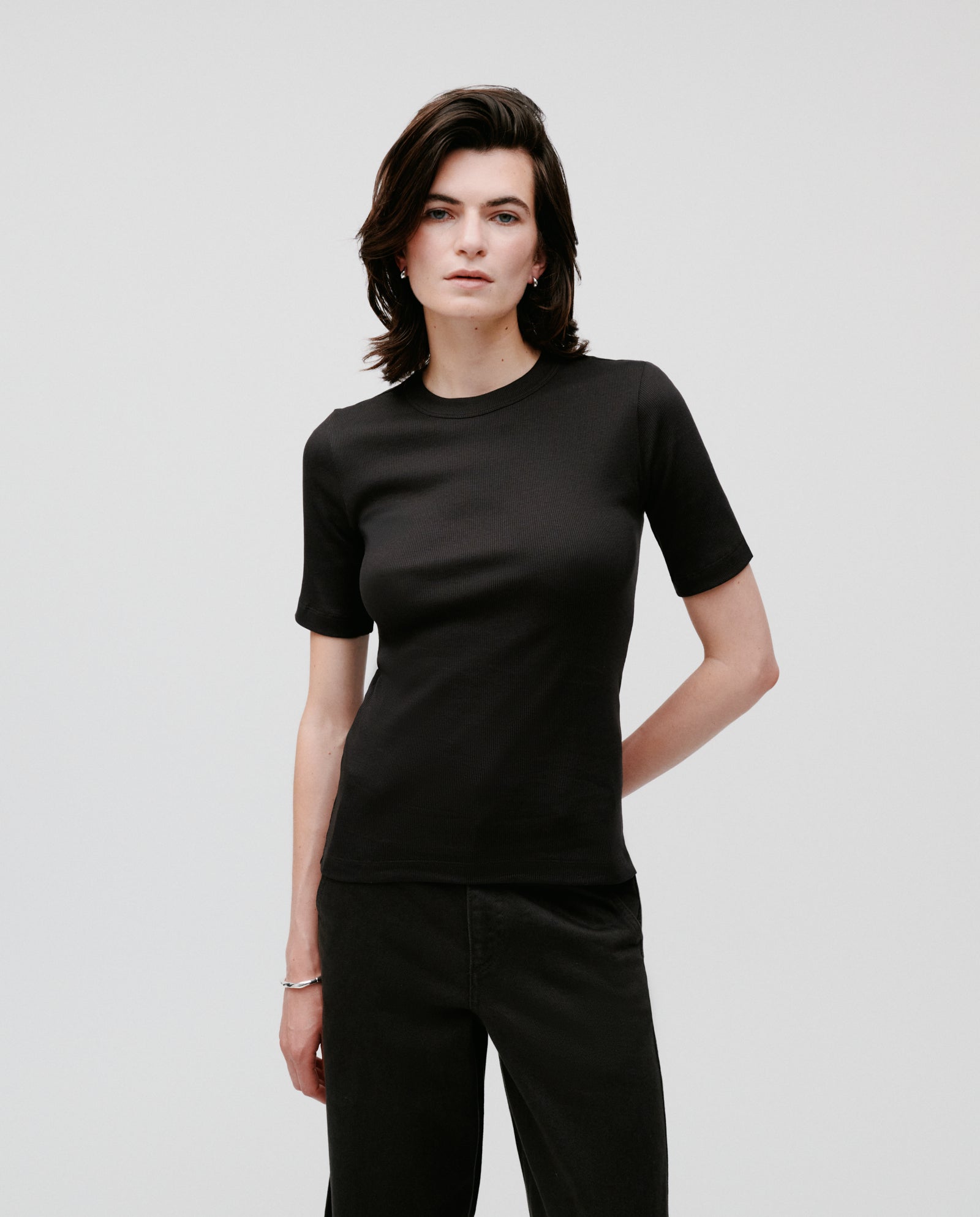 A person with short dark hair wears a fitted IVY OAK KRISTIN Top made from comfortable organic cotton and black pants, standing against a plain background.