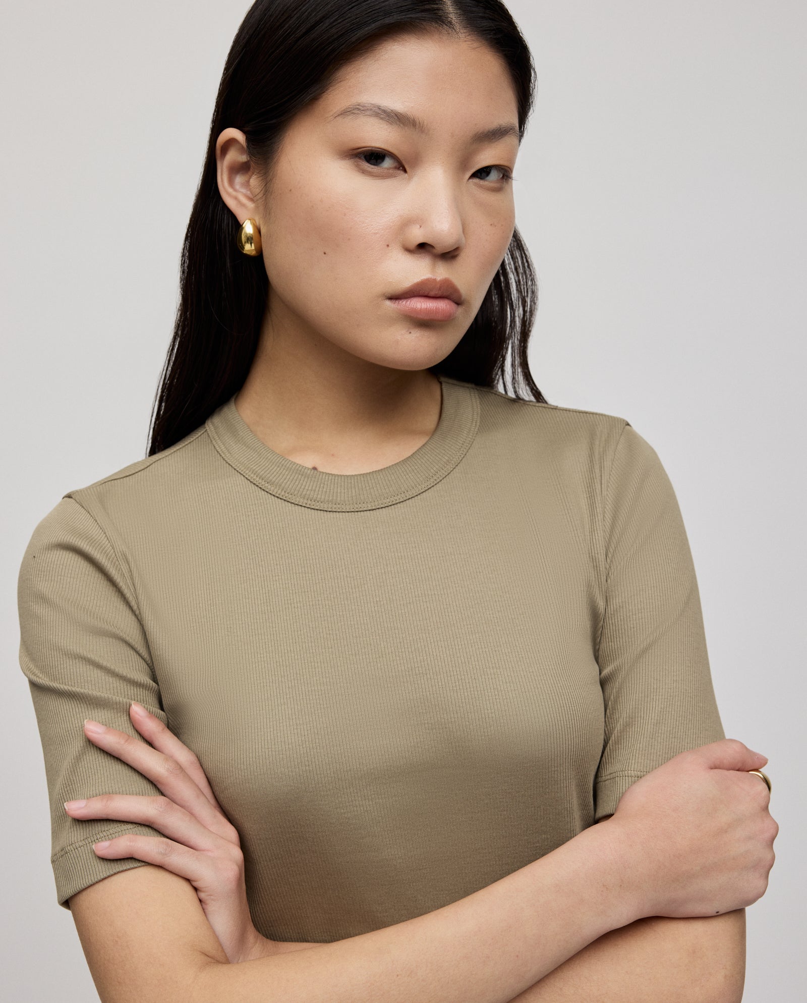 Woman with long dark hair wears a beige KRISTIN Top by IVY OAK, made from organic cotton, paired with gold hoop earrings and arms crossed—comfortable for all-day wear.