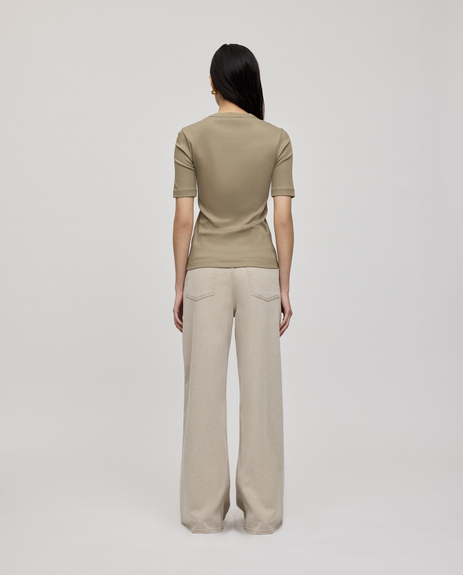 A woman stands facing away, wearing the IVY OAK KRISTIN Top in tan organic cotton with loose beige pants against a plain background—a comfortable all-day outfit.