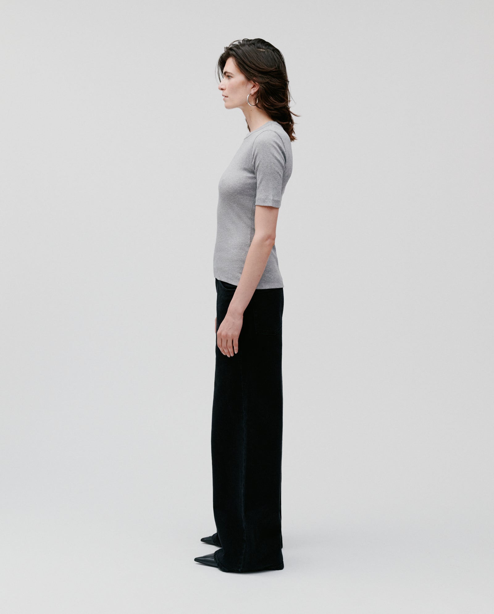 A woman stands in profile against a plain light background, wearing a comfortable gray IVY OAK KRISTIN Top made from organic cotton and black pants.