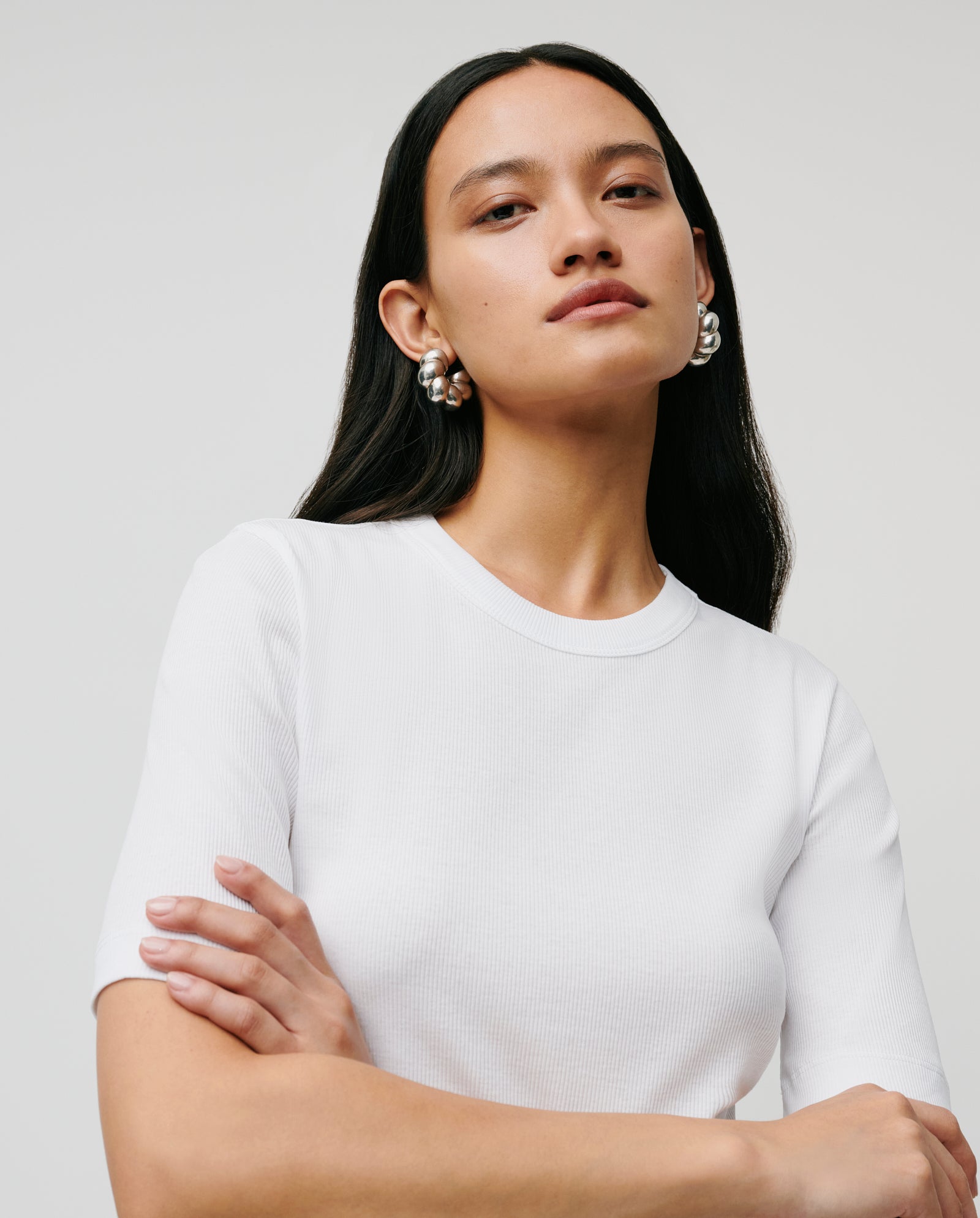Woman with long dark hair wears the comfortable IVY OAK KRISTIN Top made from organic cotton, silver earrings shining, arms crossed as she gazes ahead with confidence.