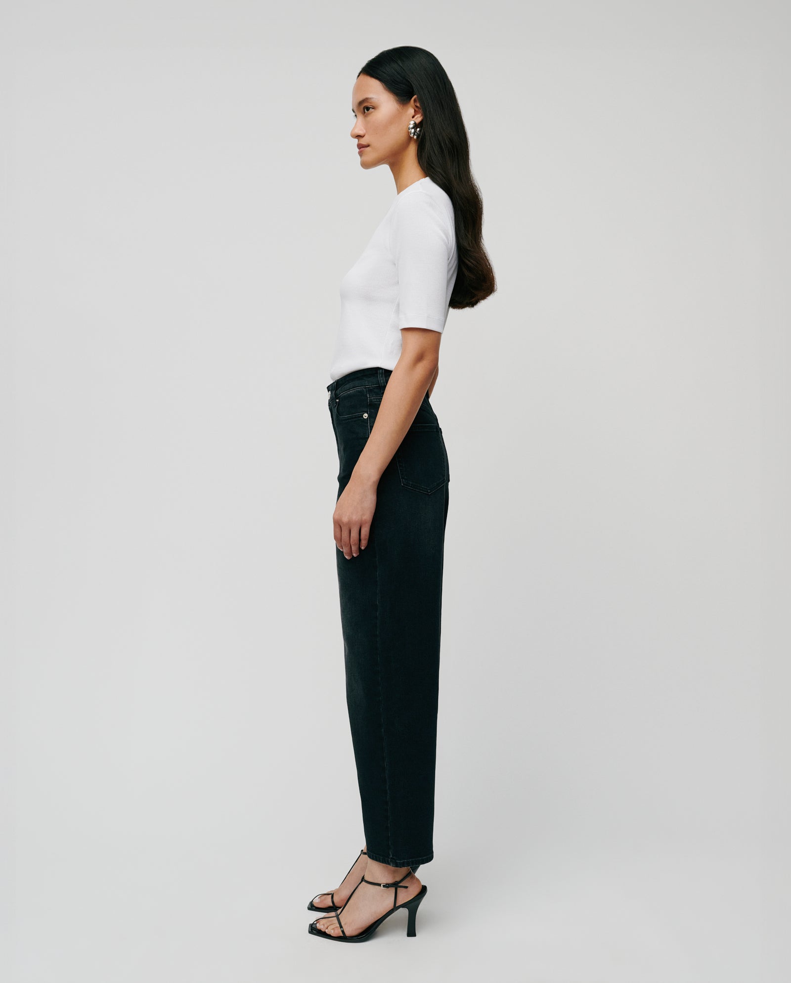 A woman in profile wears the comfortable IVY OAK KRISTIN Top in organic cotton with black jeans, black heels, and statement earrings, standing against a plain background.