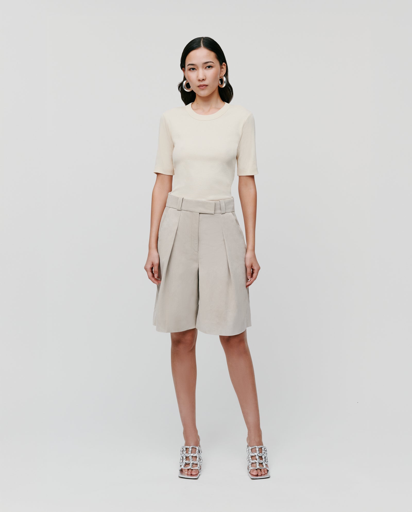 A woman wearing an IVY OAK KRISTIN Top, beige pleated shorts, and silver heels poses against a plain white background.