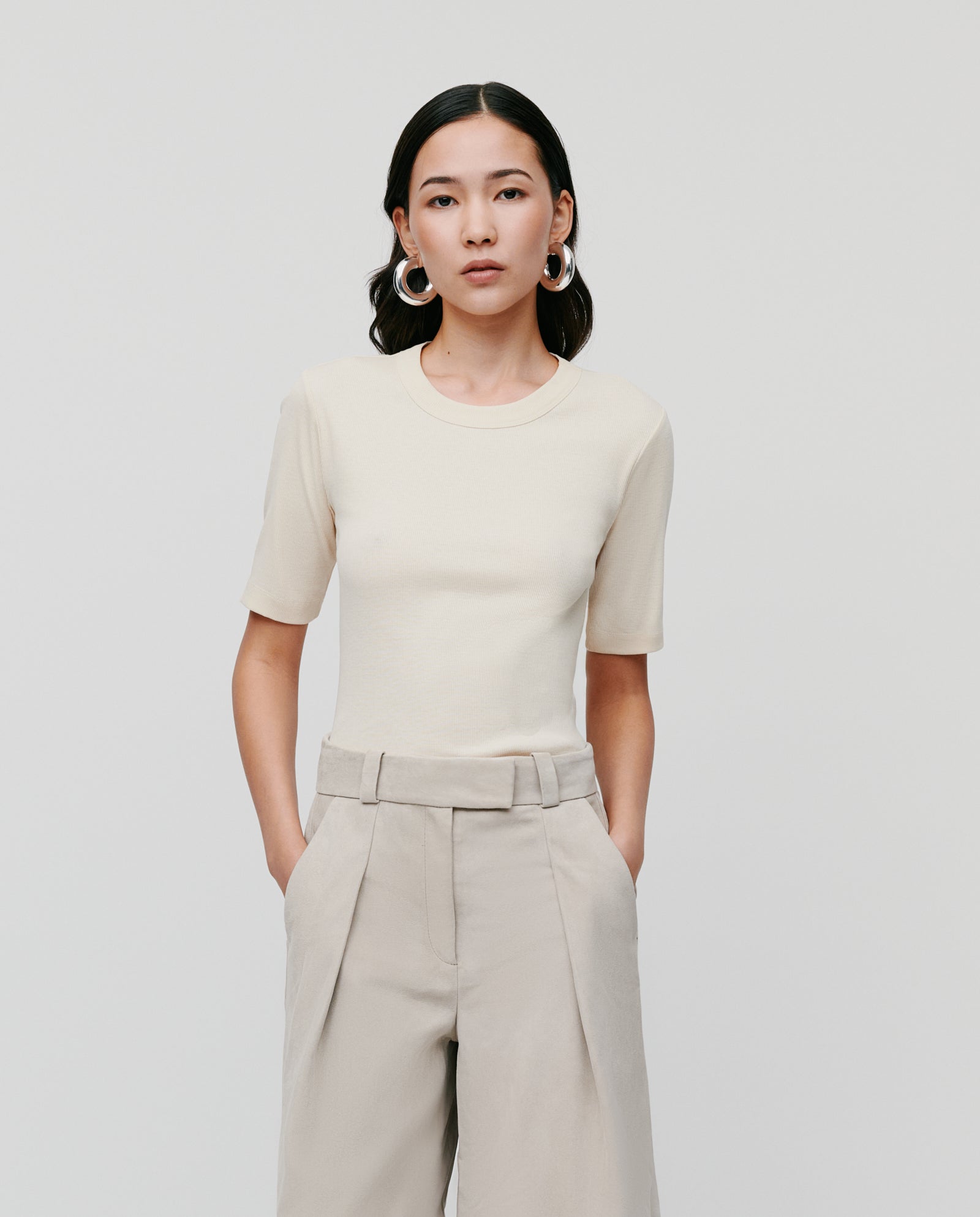 Woman in a comfortable beige KRISTIN Top by IVY OAK and high-waisted pants, hands in pockets, standing against a plain background.