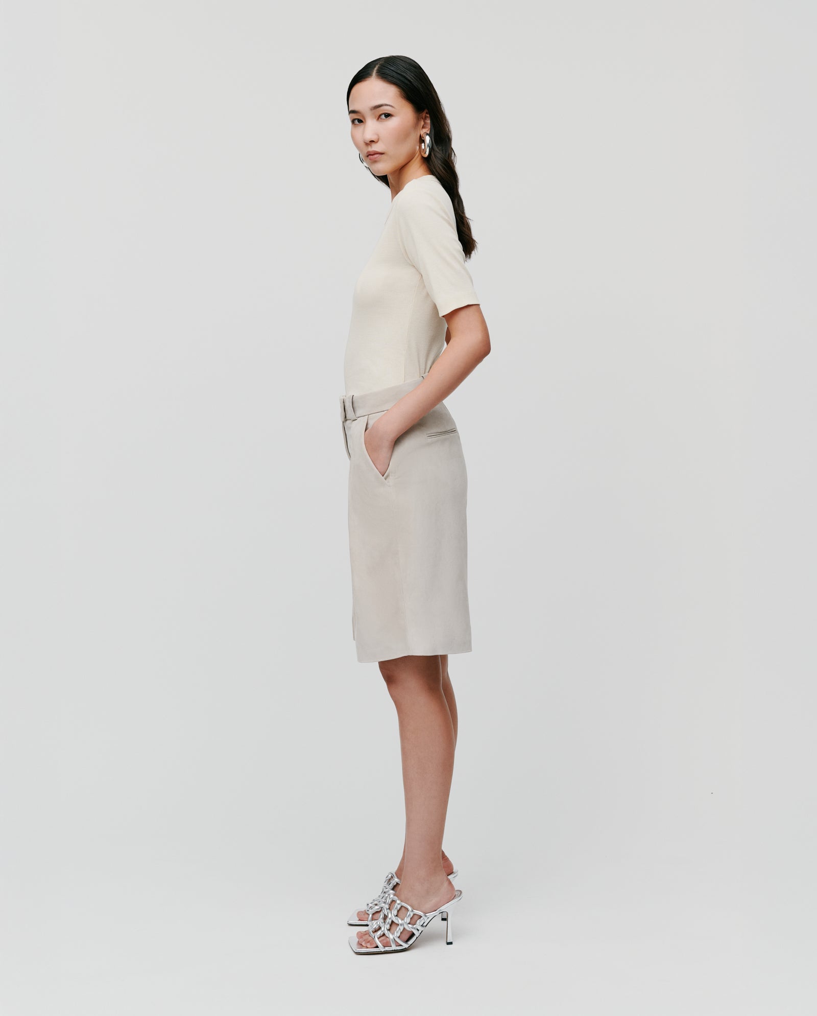 A woman stands sideways with hands in pockets, wearing the cream KRISTIN Top by IVY OAK and comfortable knee-length beige shorts against a plain background.