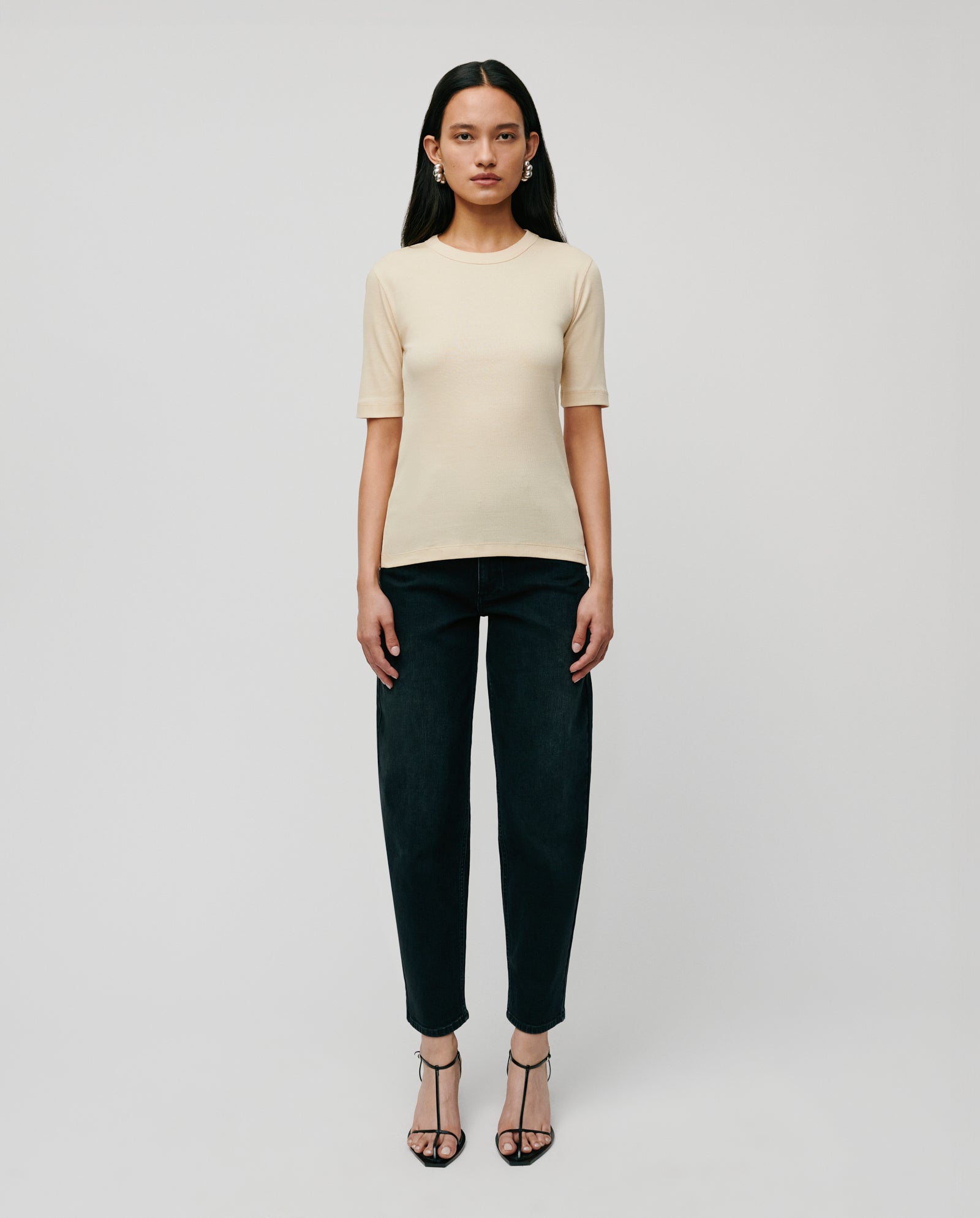 A woman stands against a plain background wearing the comfortable IVY OAK KRISTIN Top made from organic cotton, paired with black pants and black strappy heels.