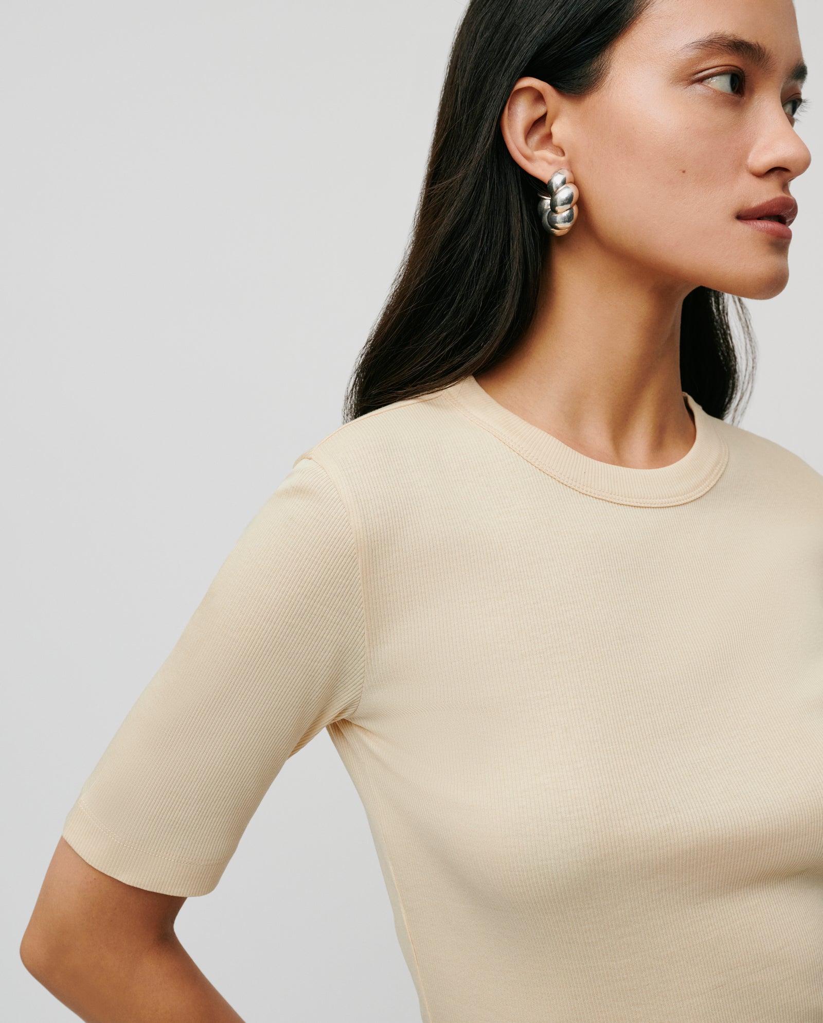 Woman in a comfortable beige KRISTIN Top by IVY OAK, crafted from organic cotton, wearing large silver hoop earrings and facing sideways.