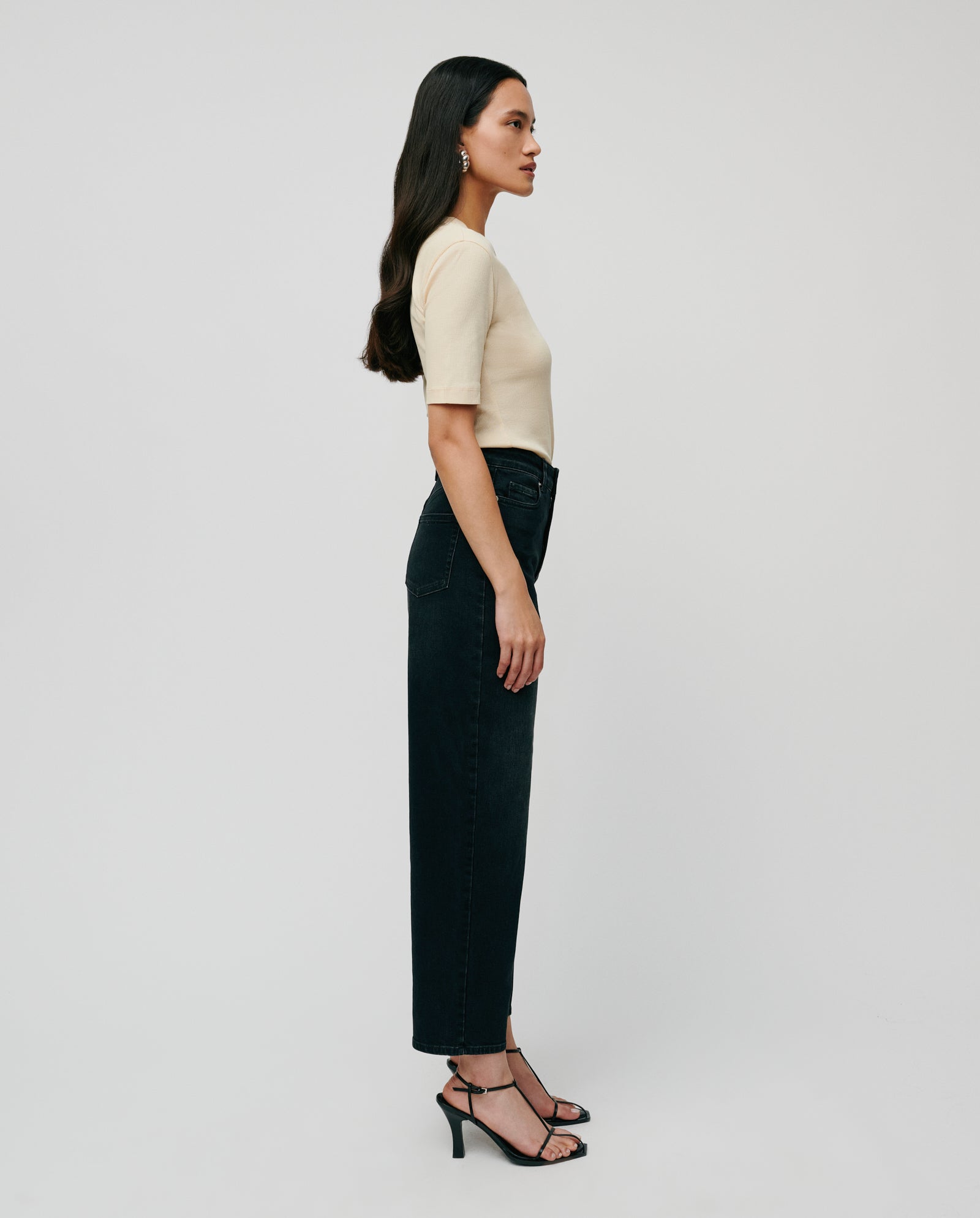 A woman in an IVY OAK KRISTIN Top, black high-waisted jeans, and black heels stands in profile against a plain background.