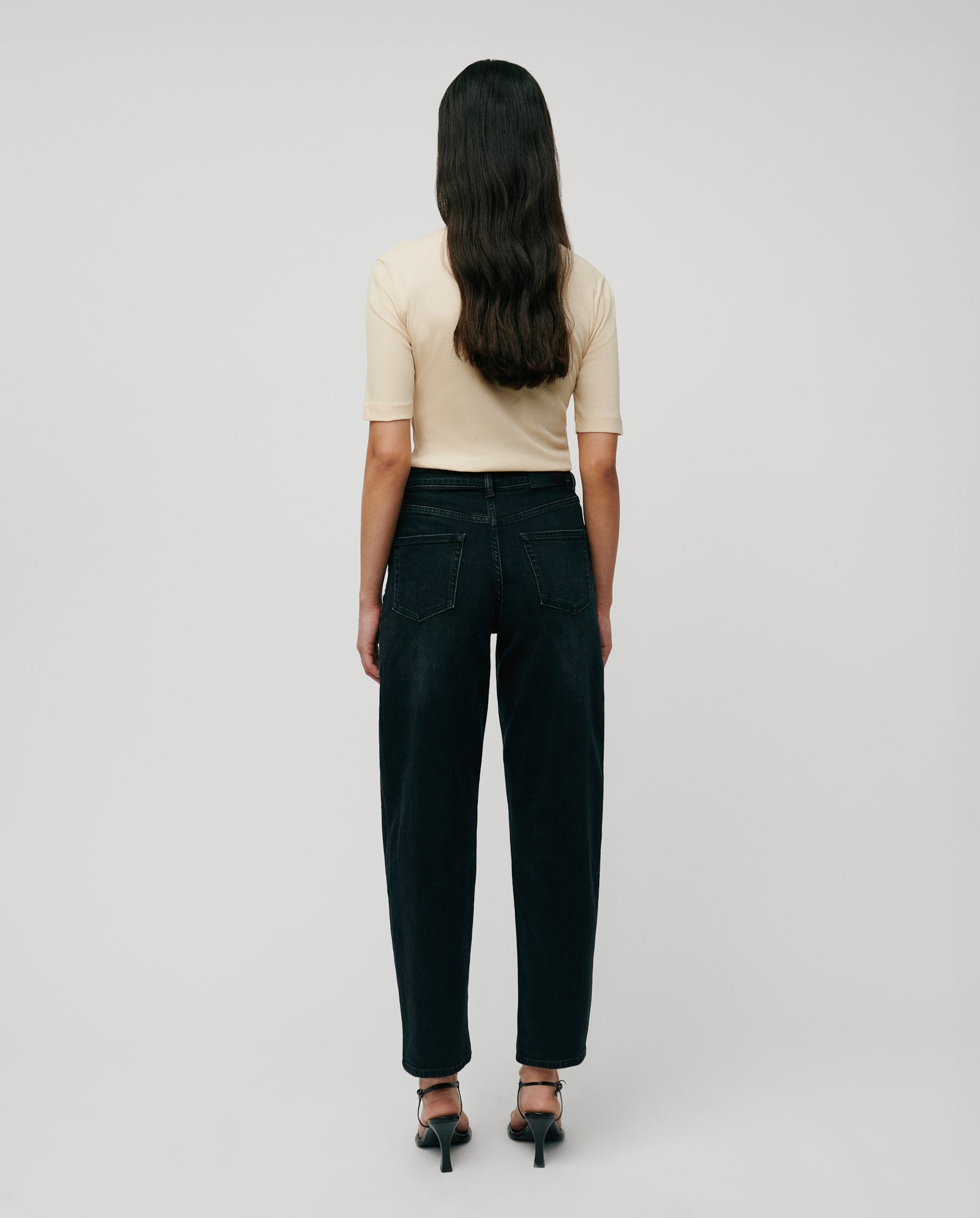 A woman faces away, wearing the comfortable beige KRISTIN Top by IVY OAK, crafted from organic cotton, styled with dark jeans and black heels against a plain background.
