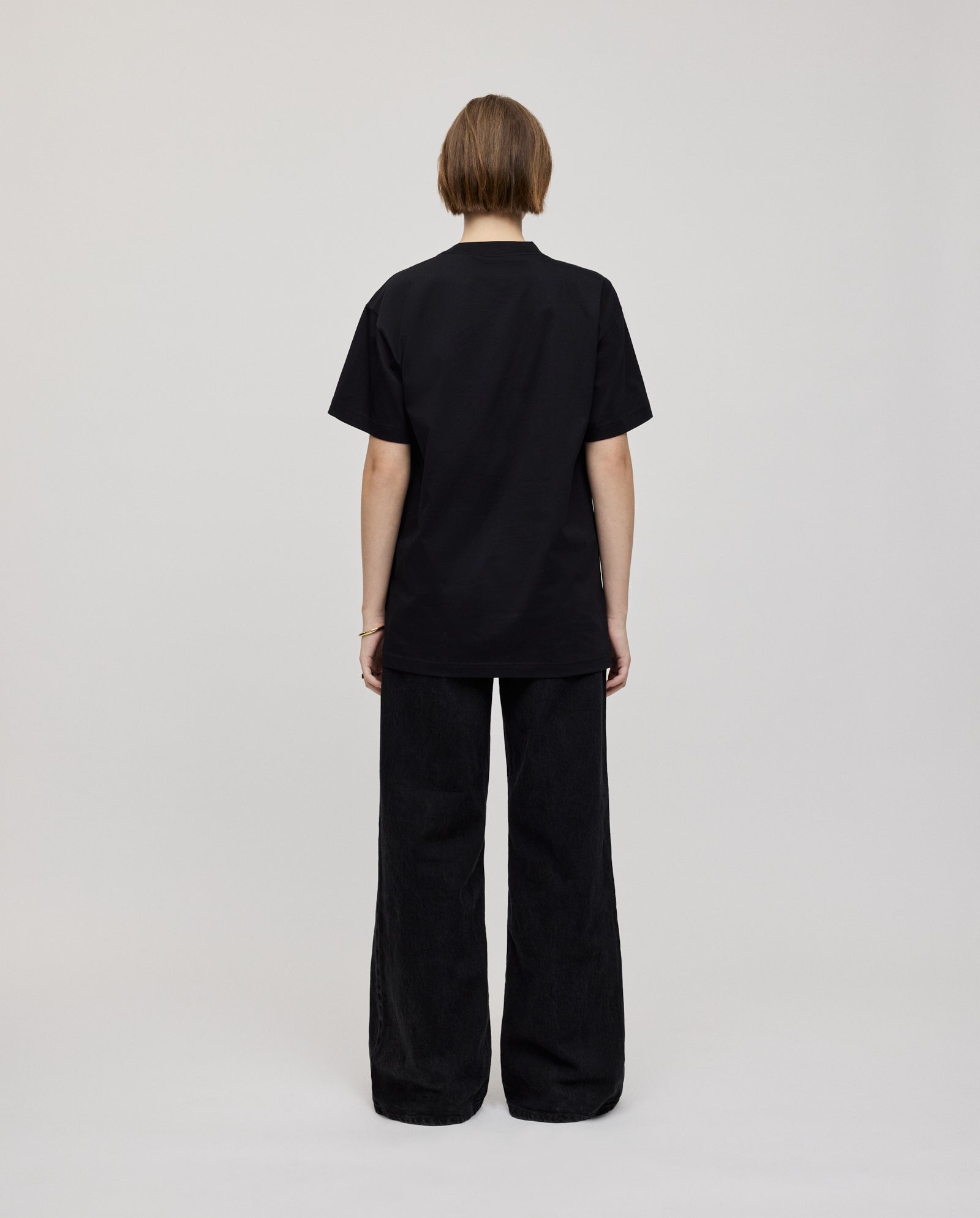 A person stands facing away against a plain background, wearing the IVY OAK KYLA ROSE Top—an ultra-soft black boxy T-shirt—paired with wide-leg black pants.