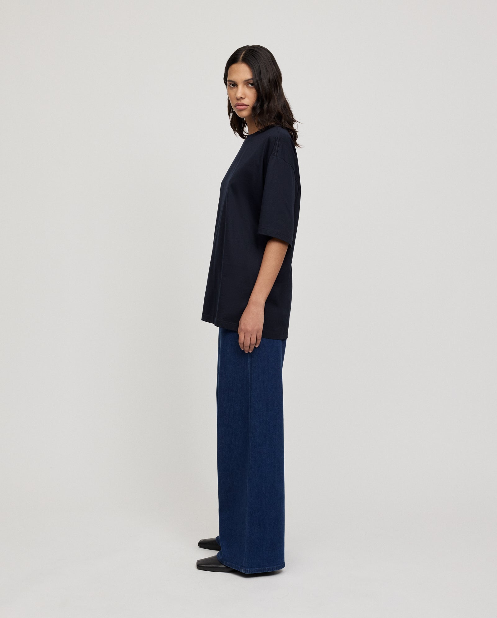 A woman stands sideways in the IVY OAK KYLA ROSE Top, styled with wide-leg blue jeans and black shoes—an ultra-soft, boxy modern essential for any wardrobe.
