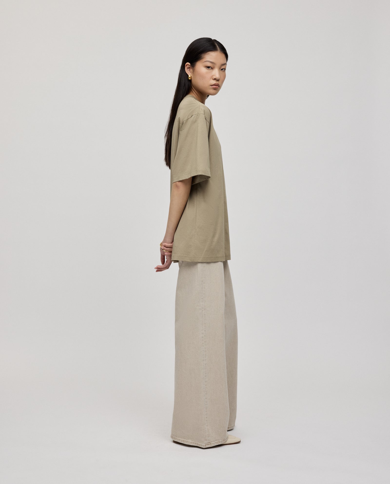 A woman stands sideways against a plain white background, wearing an ultra-soft KYLA ROSE Top by IVY OAK and wide-leg beige pants.