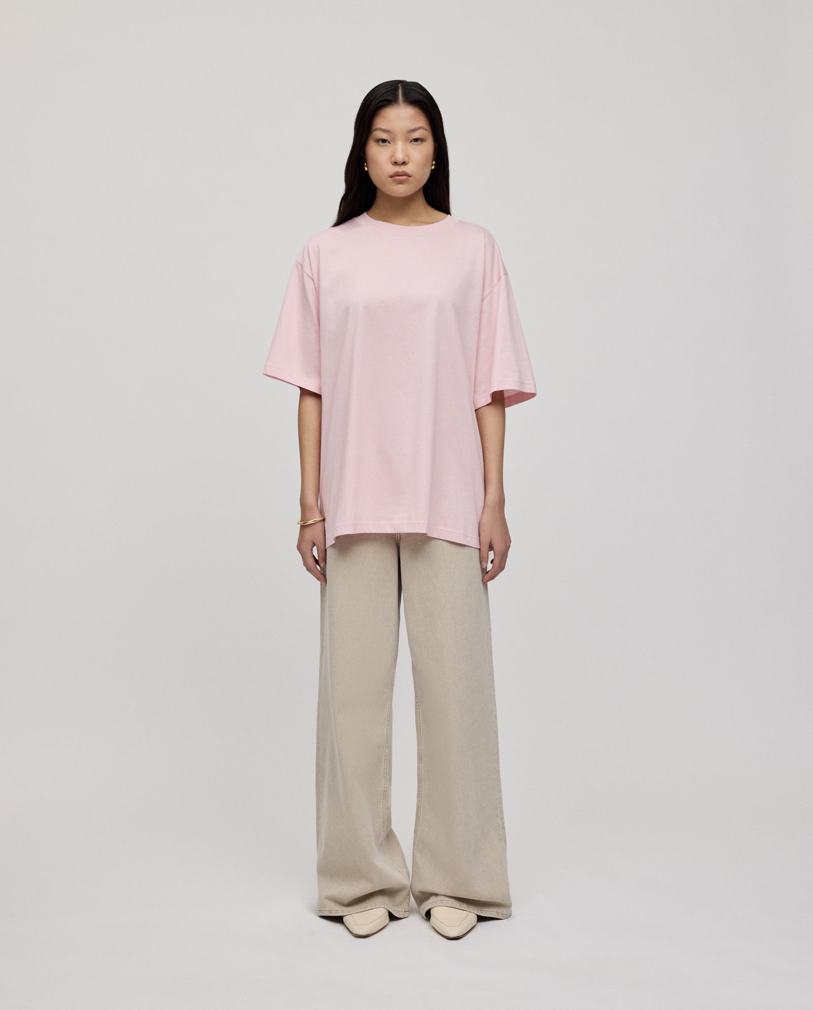 A woman stands against a plain background wearing the IVY OAK KYLA ROSE Top, an ultra-soft, light pink boxy T-shirt, paired with beige wide-leg pants.