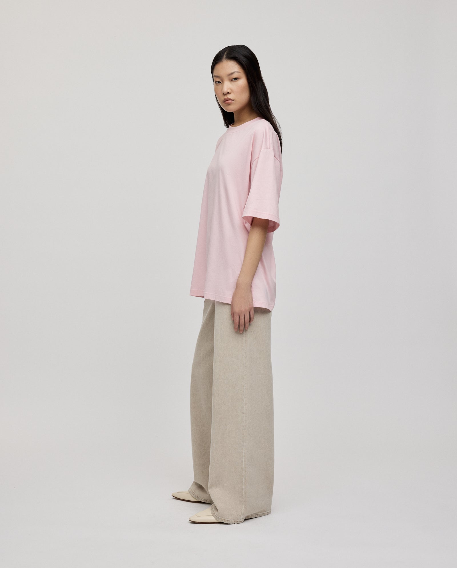 A woman stands sideways against a plain light background, wearing the IVY OAK KYLA ROSE Top, a loose fit pink ultra-soft T-shirt, paired with wide-leg beige pants.