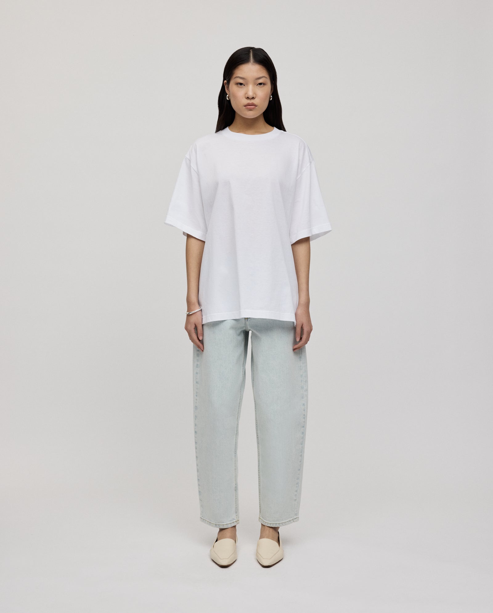 A woman faces forward wearing the IVY OAK KYLA ROSE Top in ultra-soft white, paired with light-wash jeans and cream slip-on shoes.