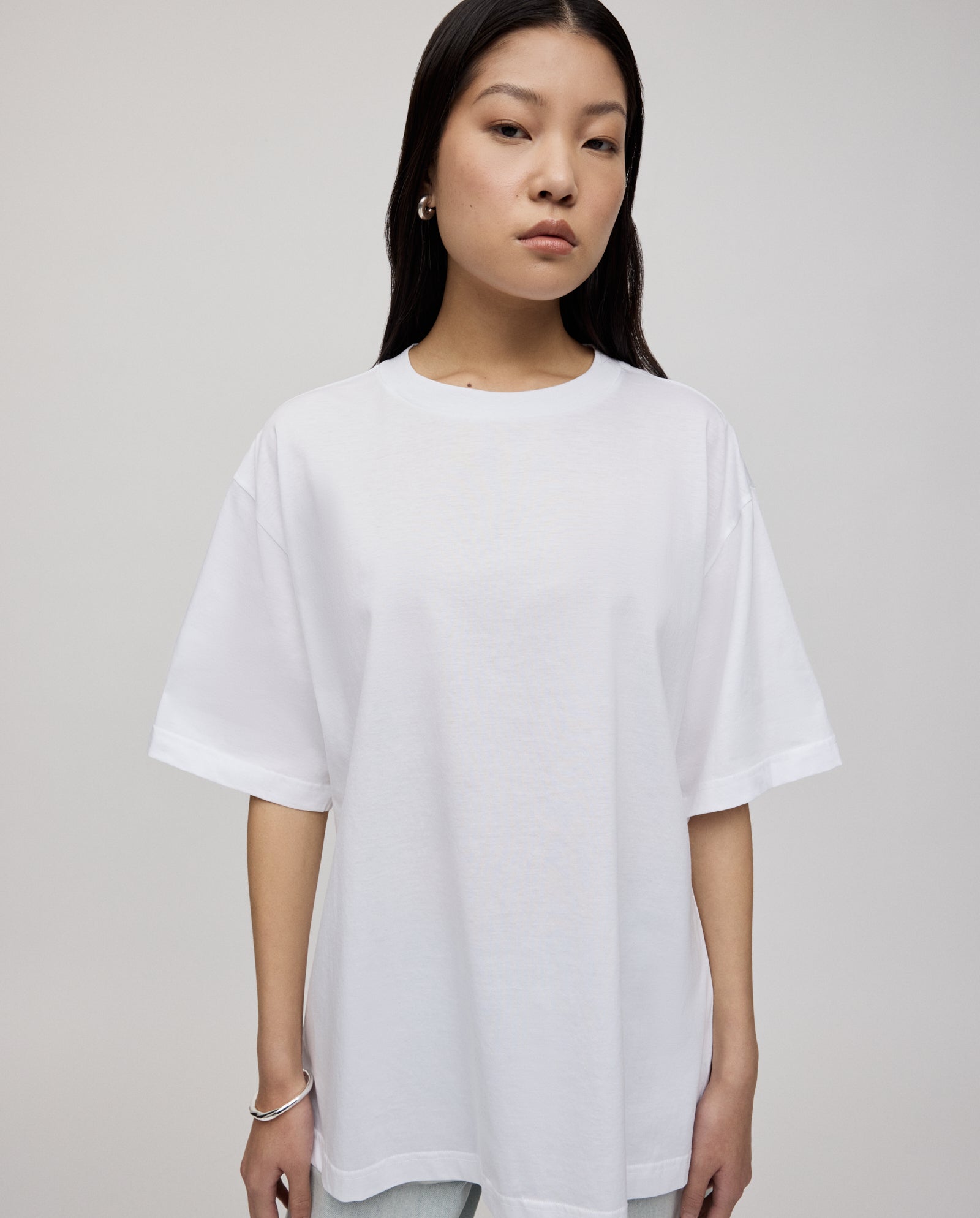 Woman with long dark hair wears the ultra-soft, loose fit KYLA ROSE Top by IVY OAK in white with light jeans, standing against a plain background.