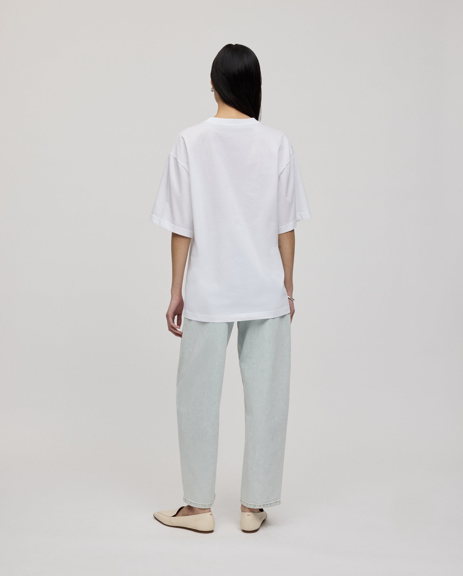 A person stands facing away against a plain background, wearing the IVY OAK KYLA ROSE Top, a loose fit white T-shirt, paired with light jeans and beige flats.