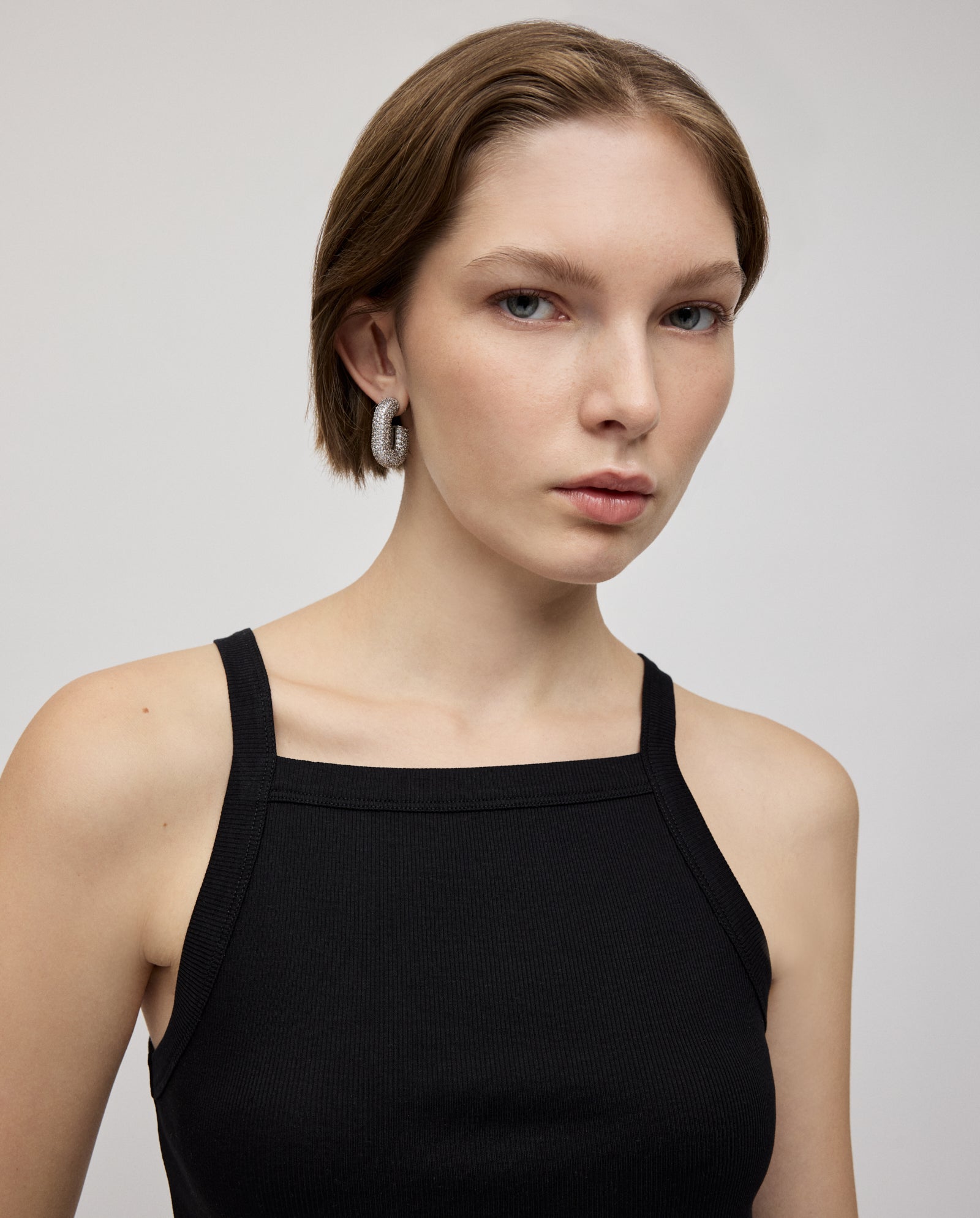 A woman with short hair and narrow shoulders models the IVY OAK KLARA ROSE Top, a black tank in ripp-optic, paired with silver hoop earrings against a light background.