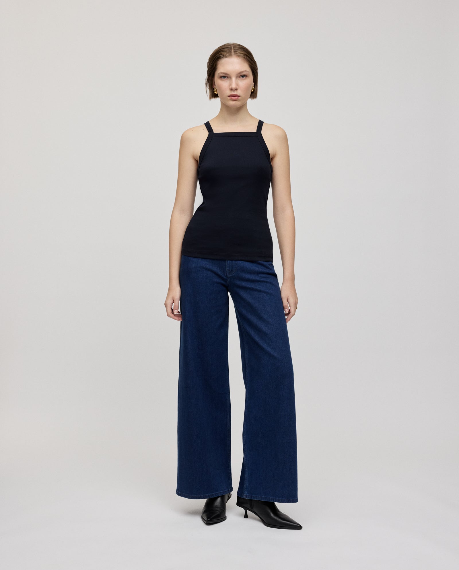 A woman with narrow shoulders wears the IVY OAK KLARA ROSE Top in black, paired with wide-leg blue jeans and black heeled boots, standing against a plain background.