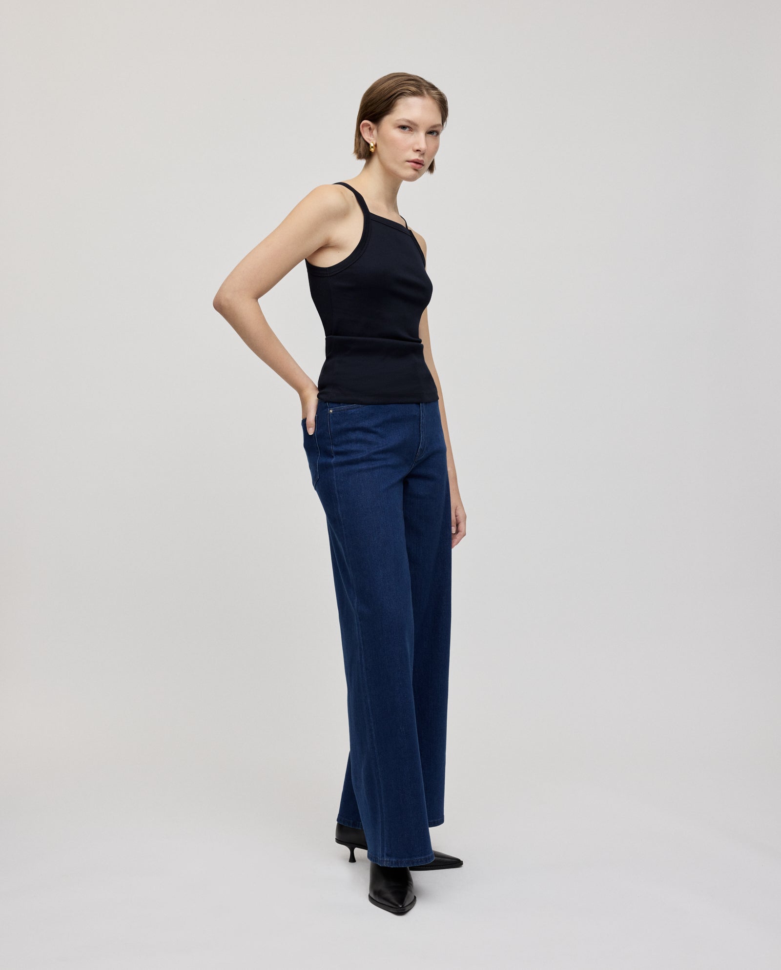 A woman wears the IVY OAK KLARA ROSE black ripp-optic tank top with narrow shoulders, paired with blue wide-leg jeans and black heels, standing against a plain background.