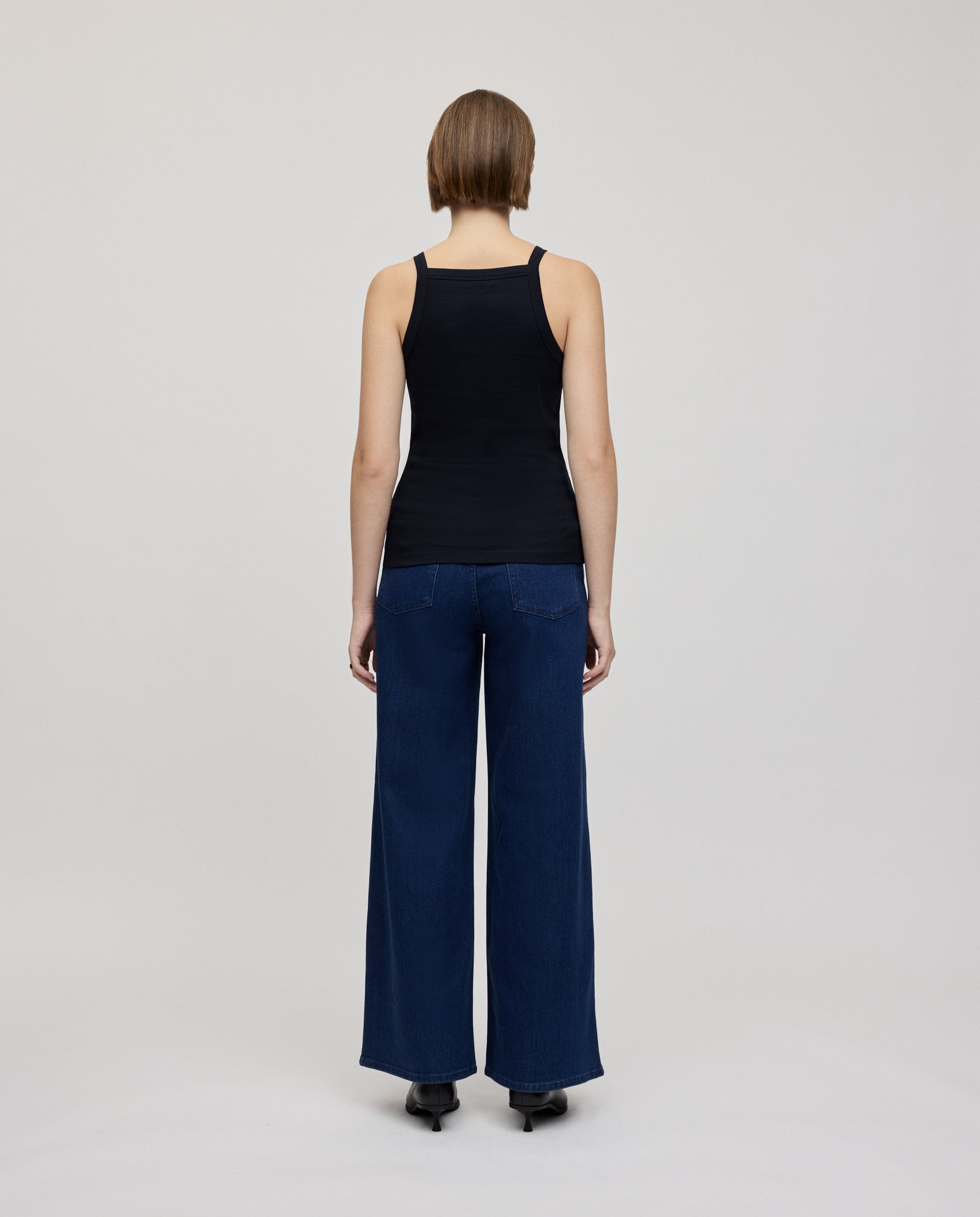 A person with narrow shoulders faces away, wearing the IVY OAK KLARA ROSE black tank top and wide-leg blue jeans against a plain background.