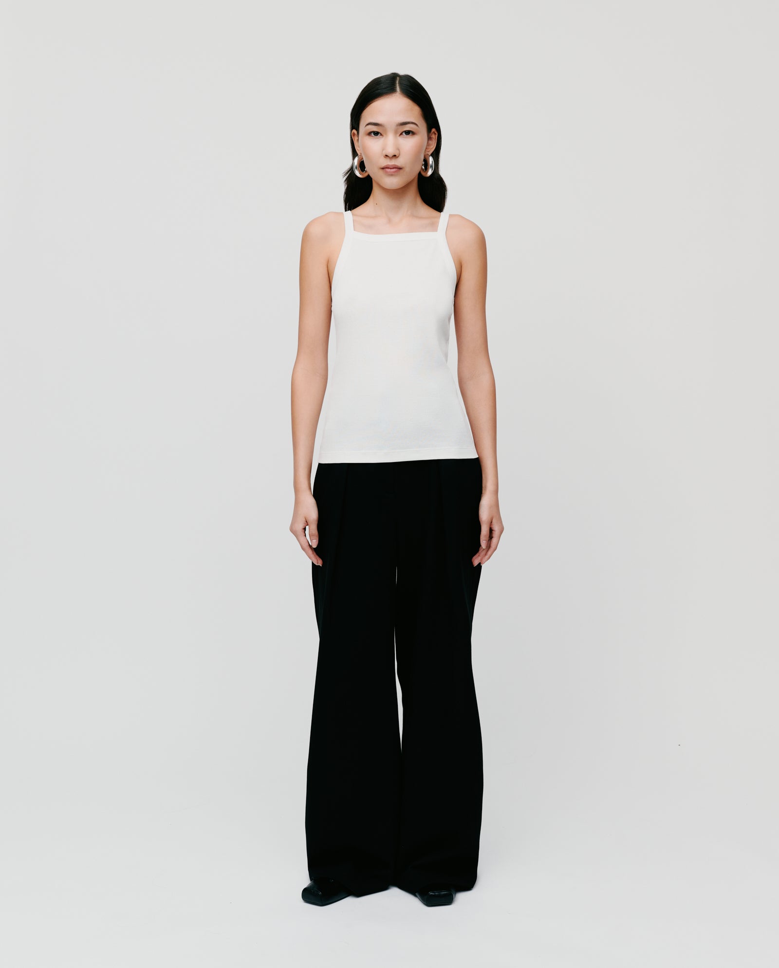 A woman with narrow shoulders stands against a plain background, wearing the IVY OAK KLARA ROSE Top in white ripp-optic and black wide-leg pants.