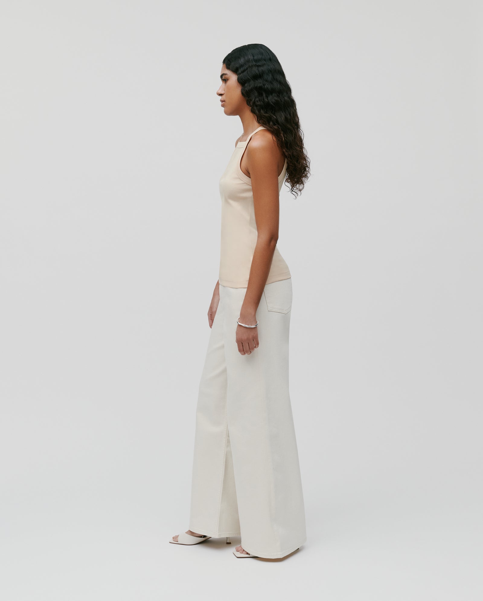 A woman stands sideways against a white background, highlighting her shoulder while wearing the KLARA ROSE Top by IVY OAK and wide-leg cream pants.