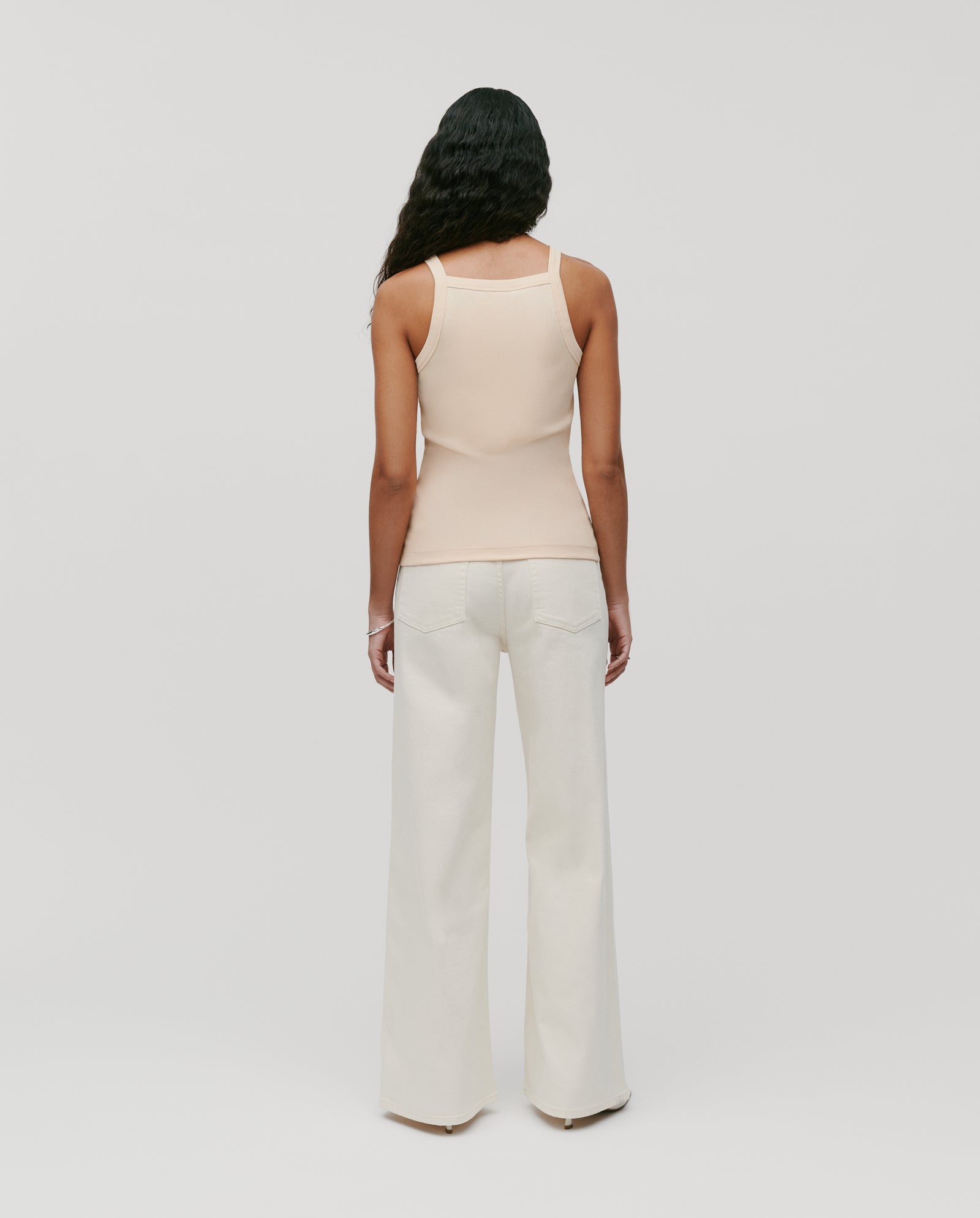 A woman with long dark hair wears the IVY OAK KLARA ROSE Top, a beige ripp-optic tank, paired with white wide-leg pants, standing turned away to highlight her shoulder.