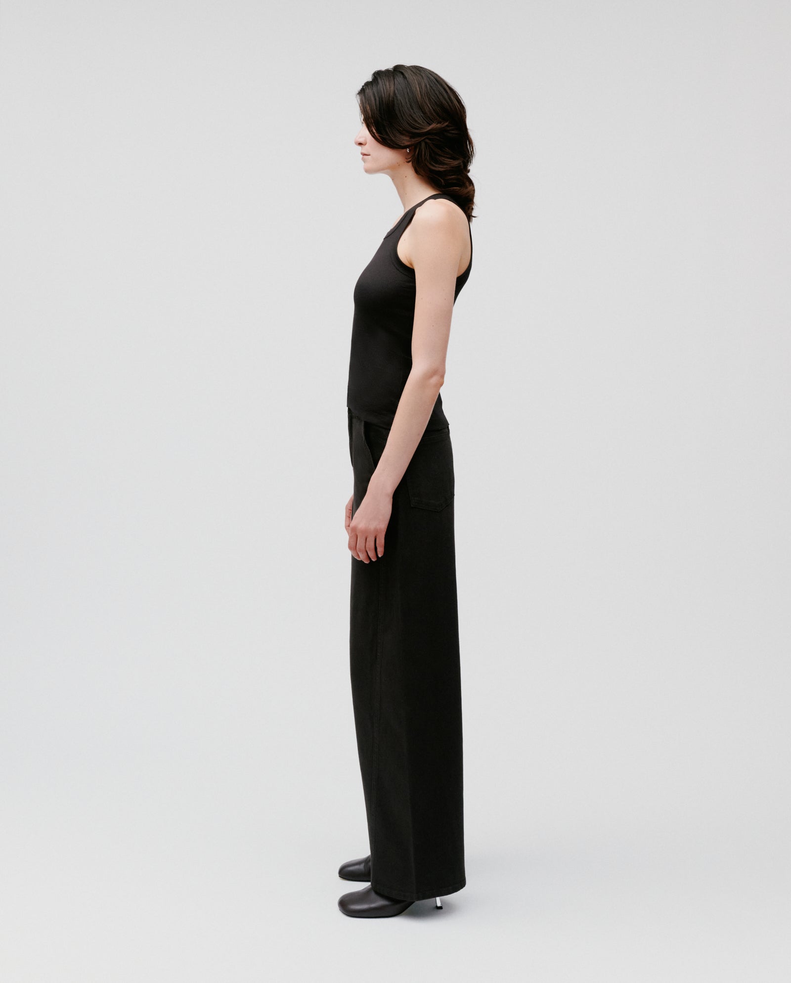 Woman wearing the IVY OAK FINLEY sleeveless black ribbed knit tank top, paired with wide-leg pants, standing sideways against a plain white background.