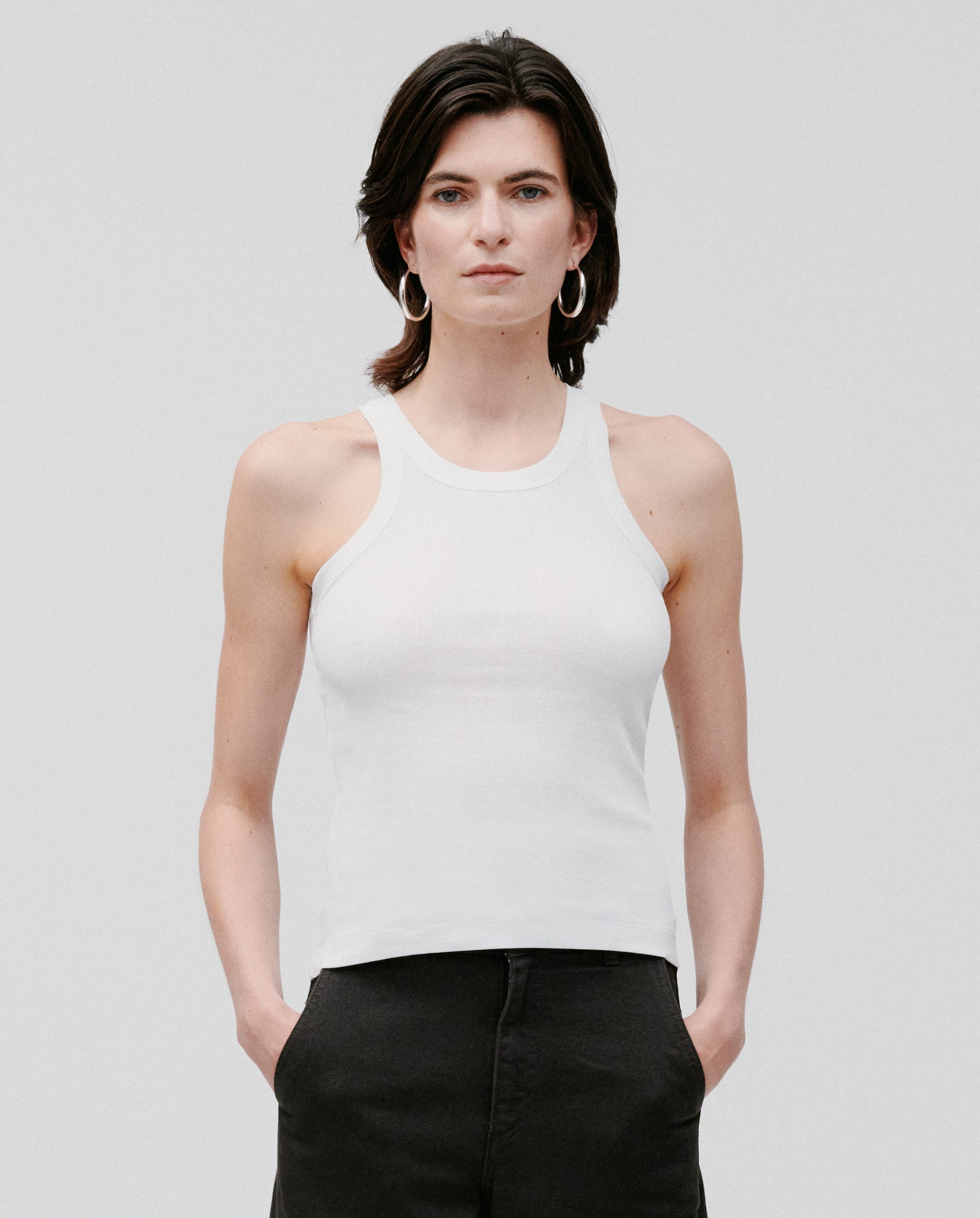 Person with short dark hair wears the white FINLEY Top by IVY OAK and black pants, standing against a plain background.