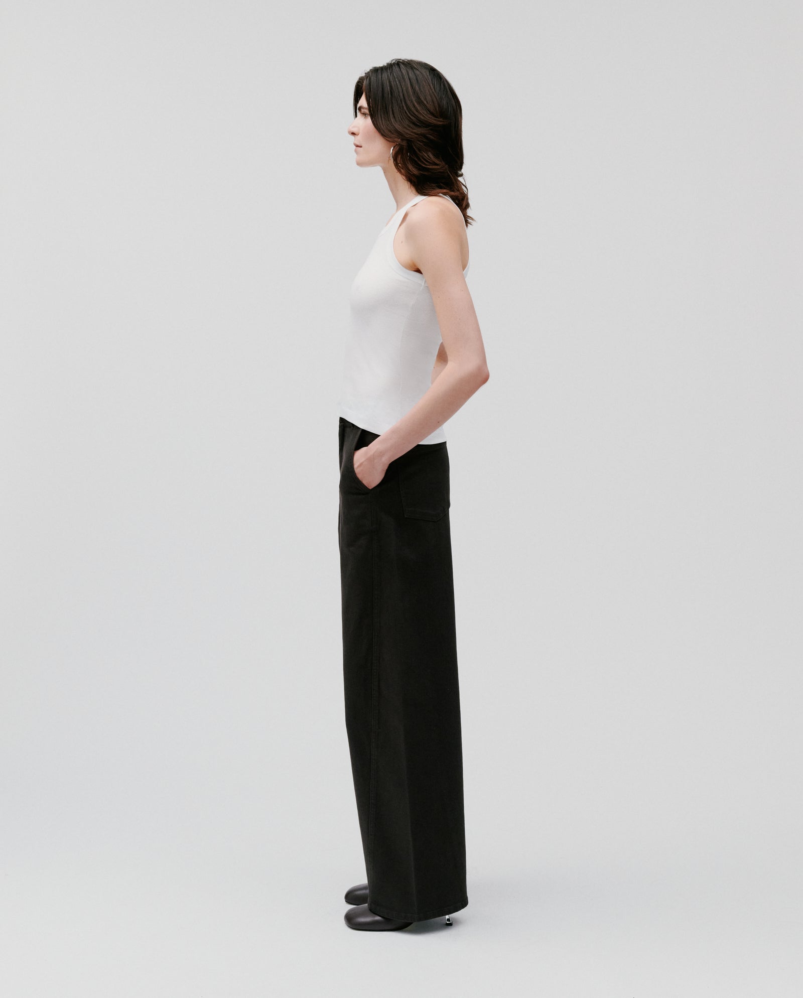 A woman stands in profile against a plain light background, wearing the IVY OAK FINLEY Top—a fine knit ribbed tank—and black wide-leg pants.