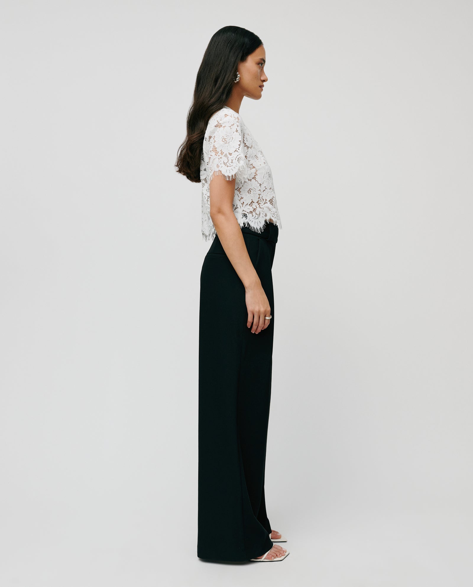 Woman in a white lace top, IVY OAK PEONY ROSE high-waist black wide leg viscose-blend trousers, and white heels stands in profile against a plain background.