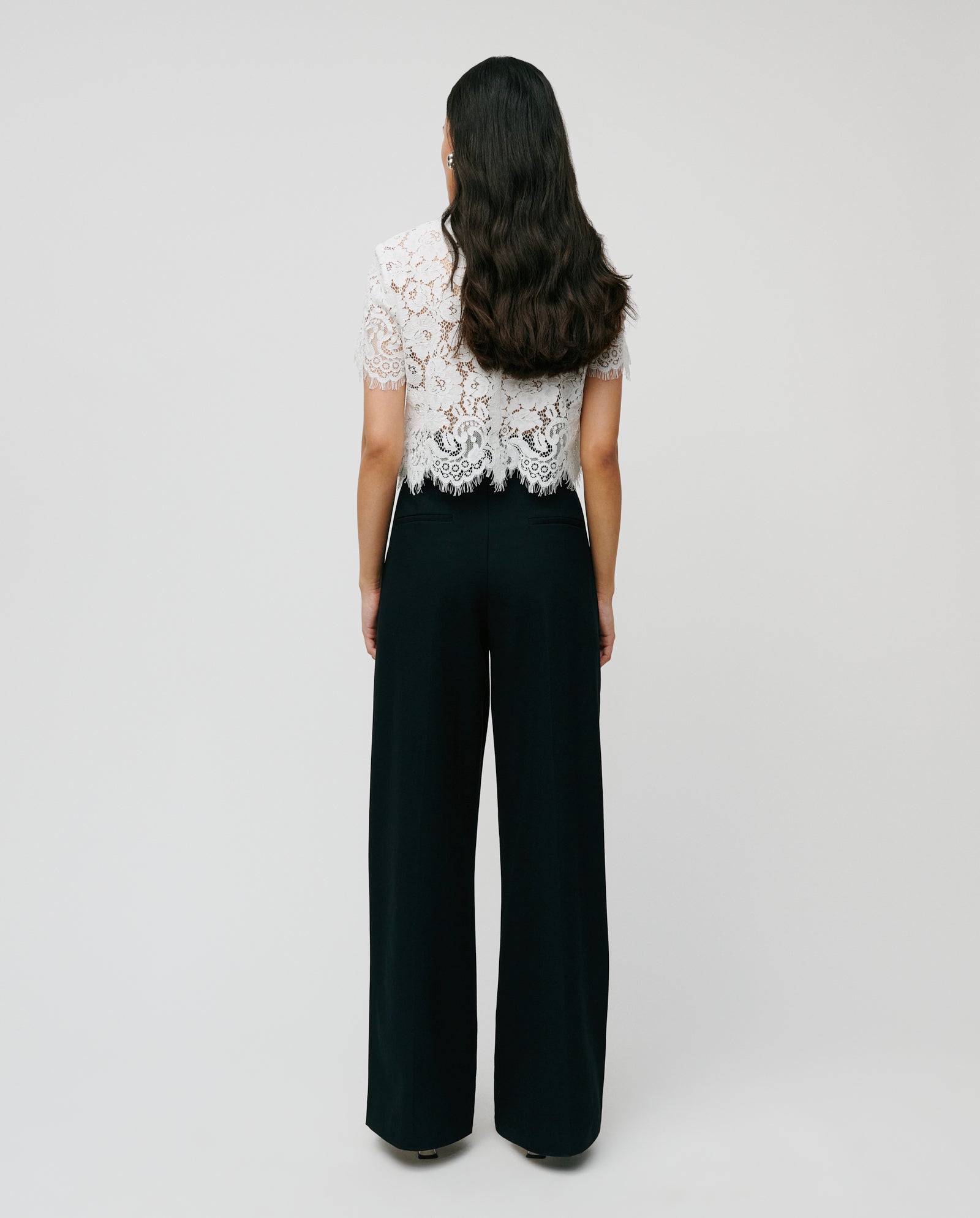 A woman faces away in a white lace top and IVY OAK PEONY ROSE high-waist black wide-leg trousers, made from a soft viscose blend, against a plain light background.
