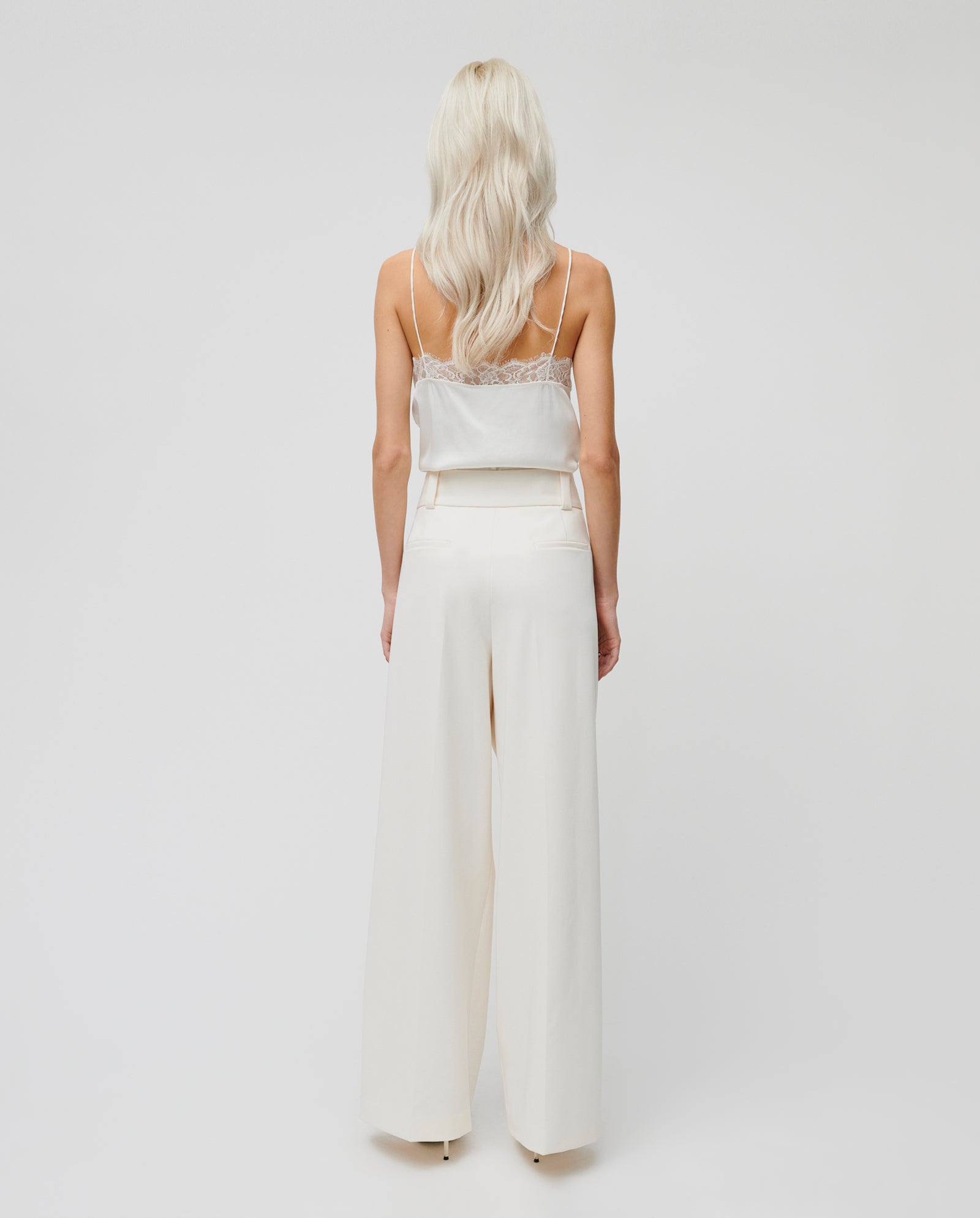 A woman with long blonde hair is seen from the back wearing IVY OAKs PEONY ROSE Trouser, a high-waisted, wide-leg pant in a soft viscose blend, paired with a white camisole.