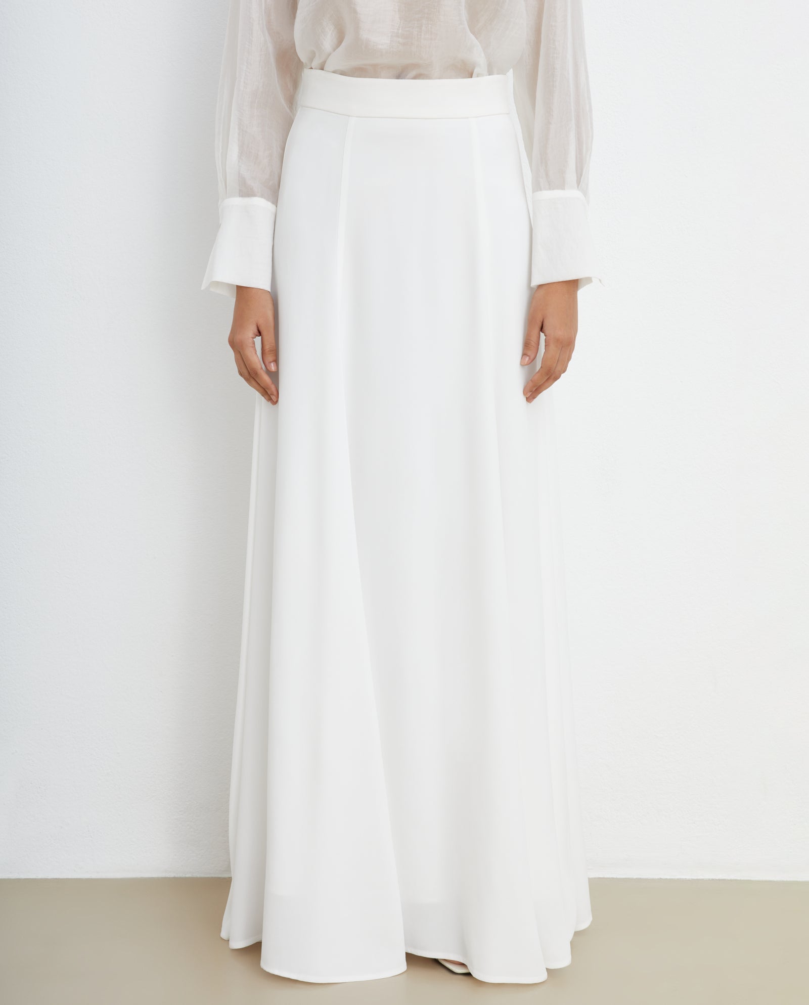 A woman stands against a white wall wearing the IVY OAK SIRENA Bridal Skirt, a long, white, high-waisted maxi skirt, paired with a sheer long-sleeve blouse.