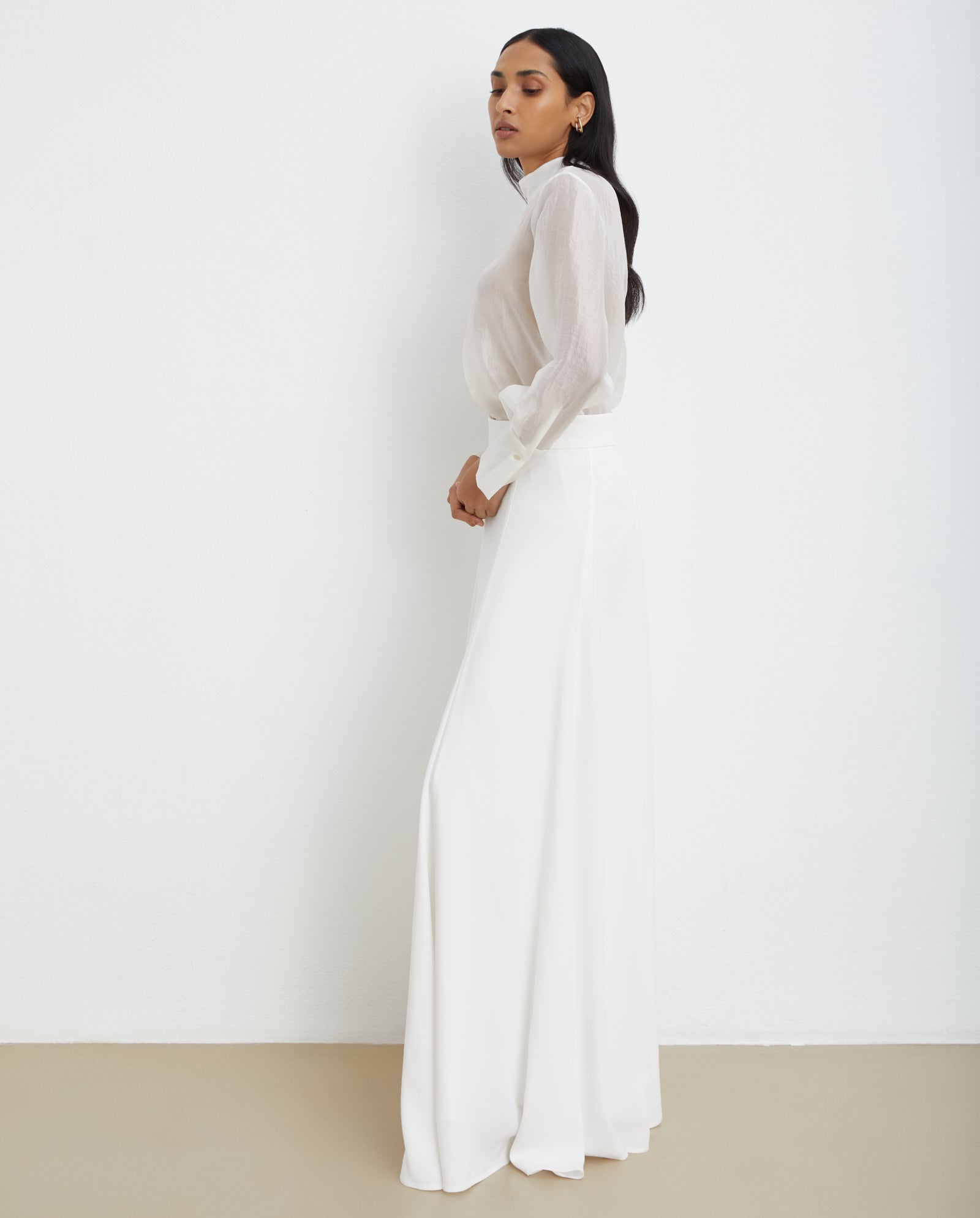 A woman in a sheer white blouse and the IVY OAK SIRENA Bridal Skirt stands sideways against a plain white wall.