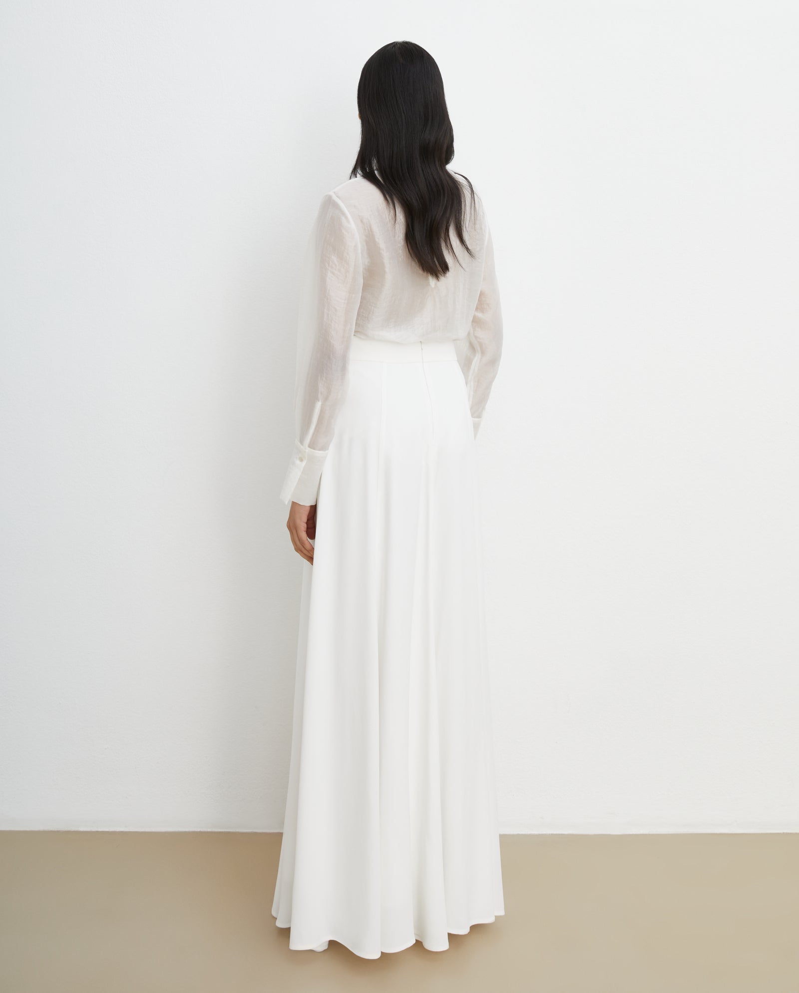 A woman in the IVY OAK SIRENA Bridal Skirt, a flowy maxi with a shiny finish, stands with her back to the camera against a white wall. Its elegant material catches the light, offering customizable lengths and styles.
