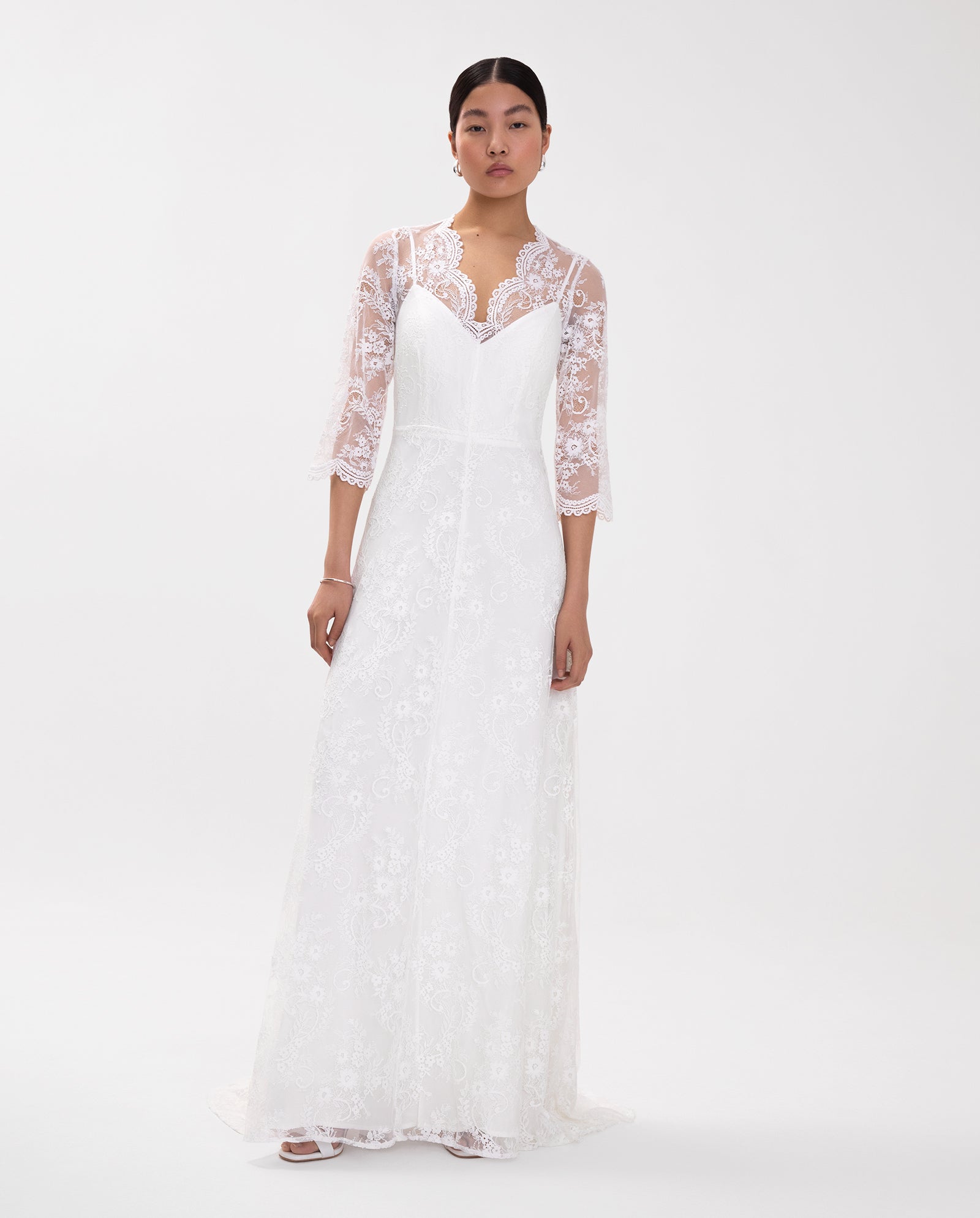 A woman wears the IVY OAK DALIA Bridal Dress—a long white lace gown with a V-neckline and sheer lace sleeves—posing against a plain white background.