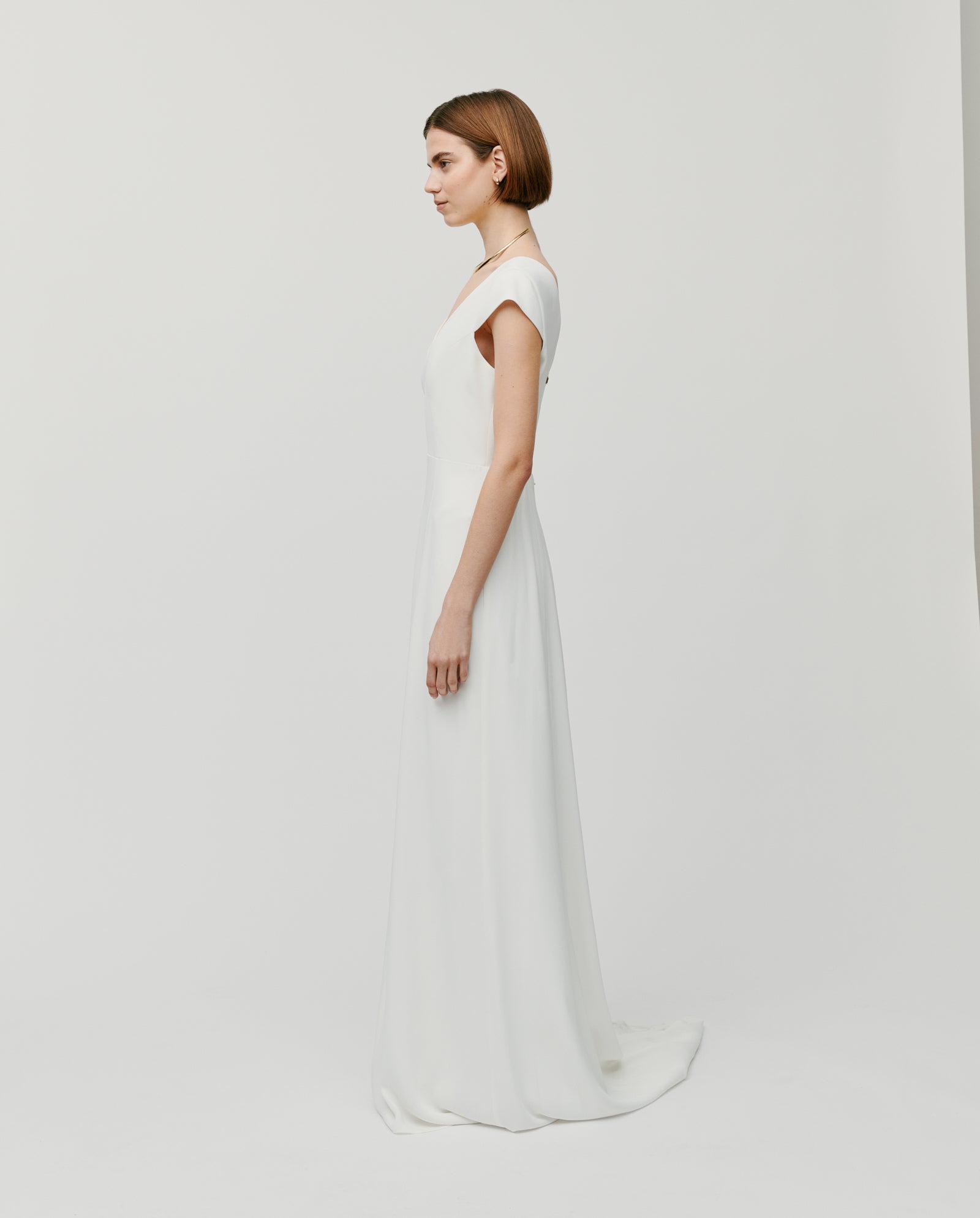 A woman in a simple, elegant white DARIA Bridal Dress by IVY OAK with a V-neckline stands sideways against a plain white background—a timeless maxi wedding dress offered in customizable lengths.