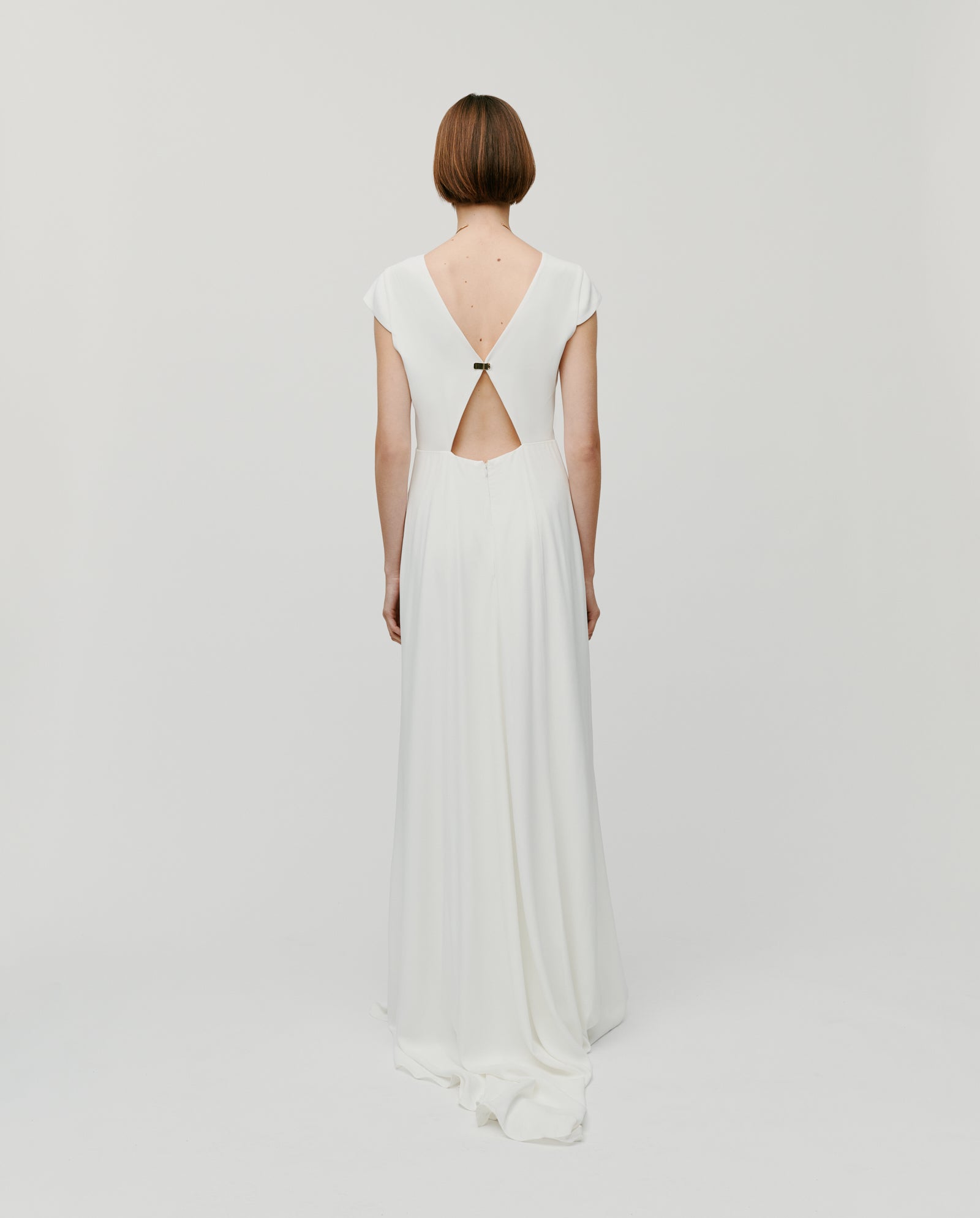 A woman wears the IVY OAK DARIA Bridal Dress, a white maxi wedding gown with cap sleeves, V-neckline, and deep V-back, standing against a light background. Customizable lengths ensure the perfect fit.