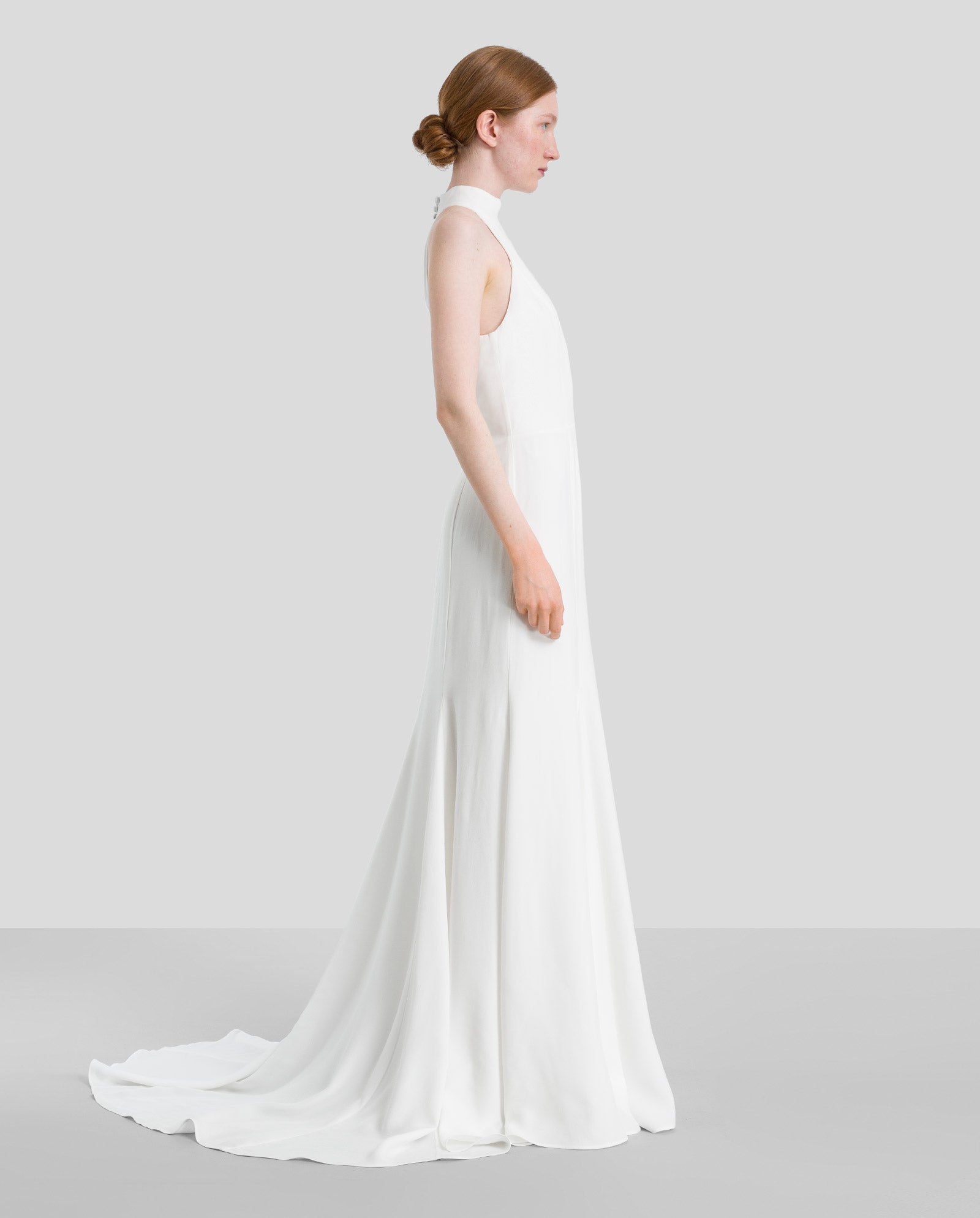 A woman in the IVY OAK MEREDITH Bridaldress, a sleeveless white American shoulder dress with subtle godets, stands in profile against a plain gray background.