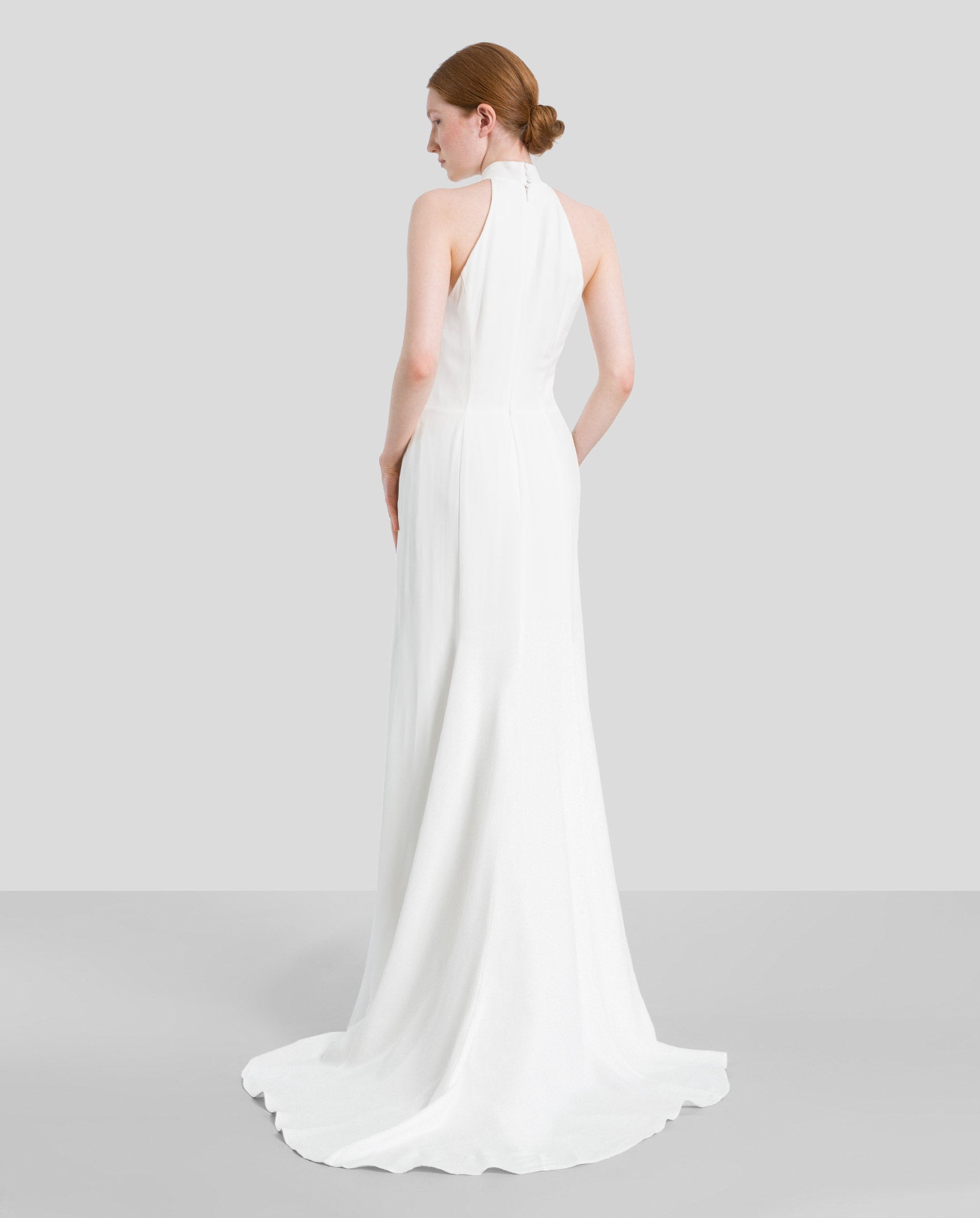 A woman stands facing away against a plain background, wearing the IVY OAK MEREDITH Bridaldress—an American shoulder wedding gown with godets and a long train.