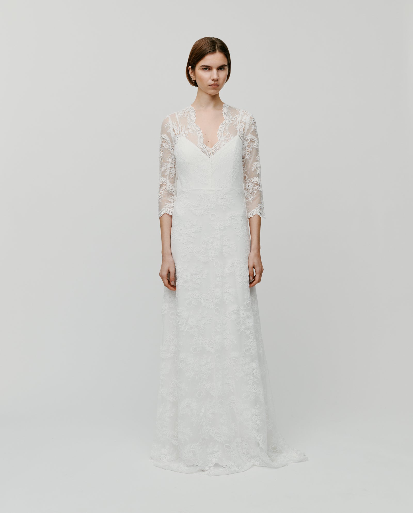 A woman models the IVY OAK DALIA AMY Dress, a long white lace gown with three-quarter sleeves and a V-neckline, standing against a plain background.