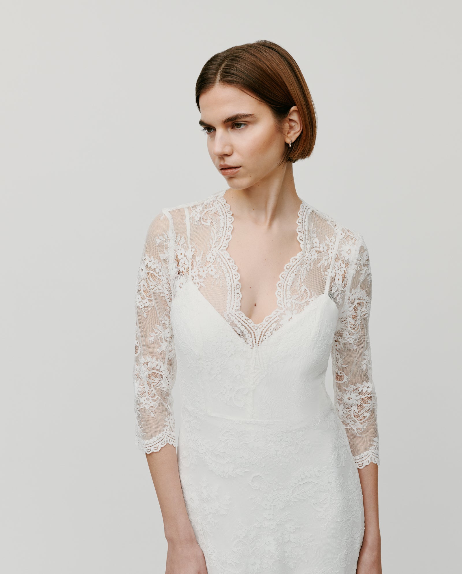 A woman wearing the IVY OAK DALIA AMY bridal lace dress, featuring sheer sleeves and a V-neckline, poses against a plain background.