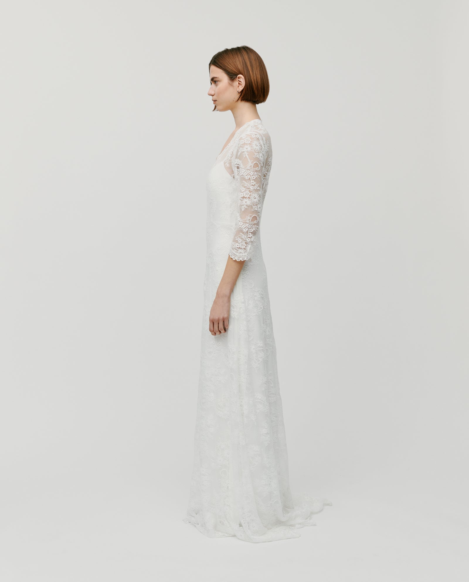 Woman in the DALIA AMY dress by IVY OAK, featuring bridal lace, three-quarter sleeves, and a V-neckline, standing in profile against a plain background.