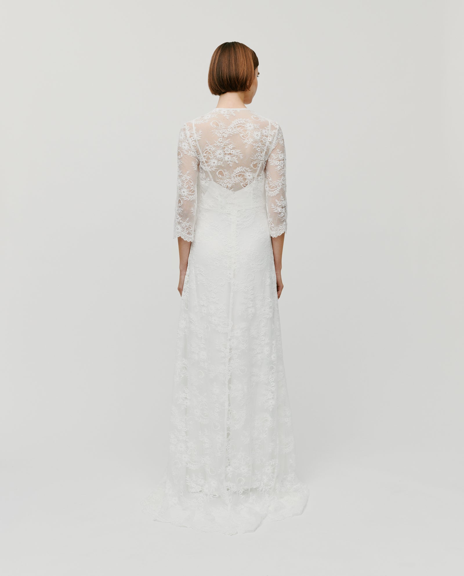 A woman stands facing away against a plain white background, wearing the IVY OAK DALIA AMY Dress—a long-sleeved bridal lace gown with a V-neckline and a skirt with train.